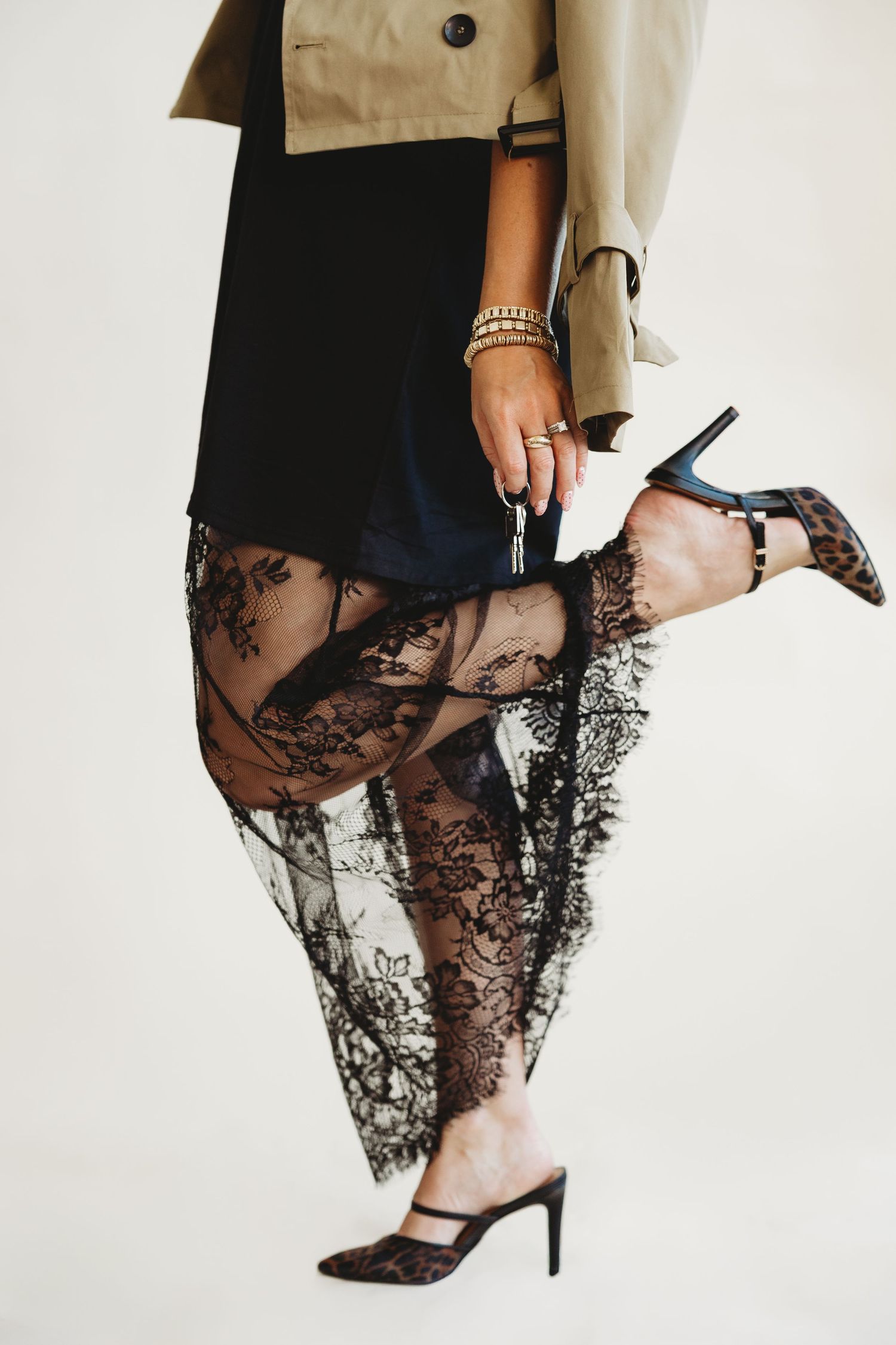 Woman in a tan trench jacket, black dress and sheer black lace skirt lifting a leg to show leopard print heels.