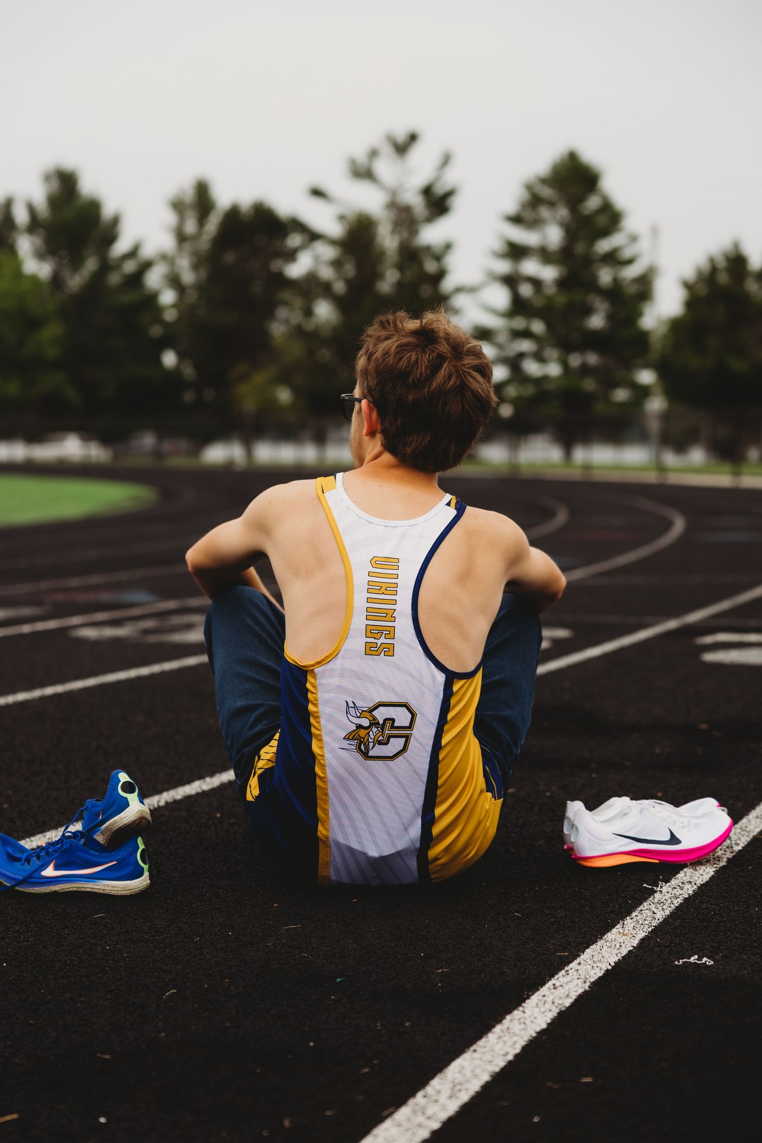 Track athlete in a white and gold jersey sits on a running track beside blue and pink running shoes, facing grass and evergreen trees.