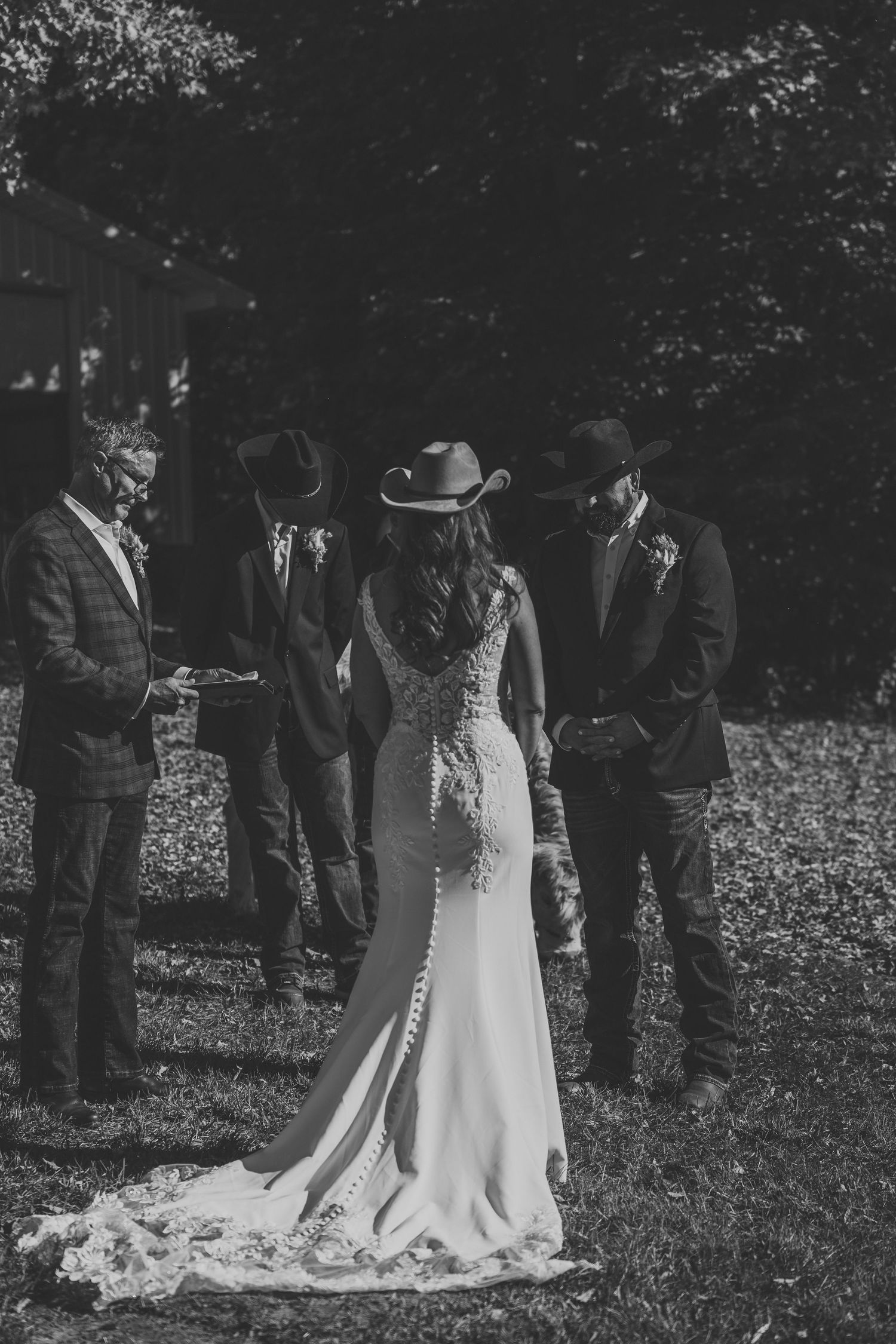 Black and white photo of a bride in a lace gown and cowboy hat at an outdoor wedding ceremony with an officiant and two men in hats.