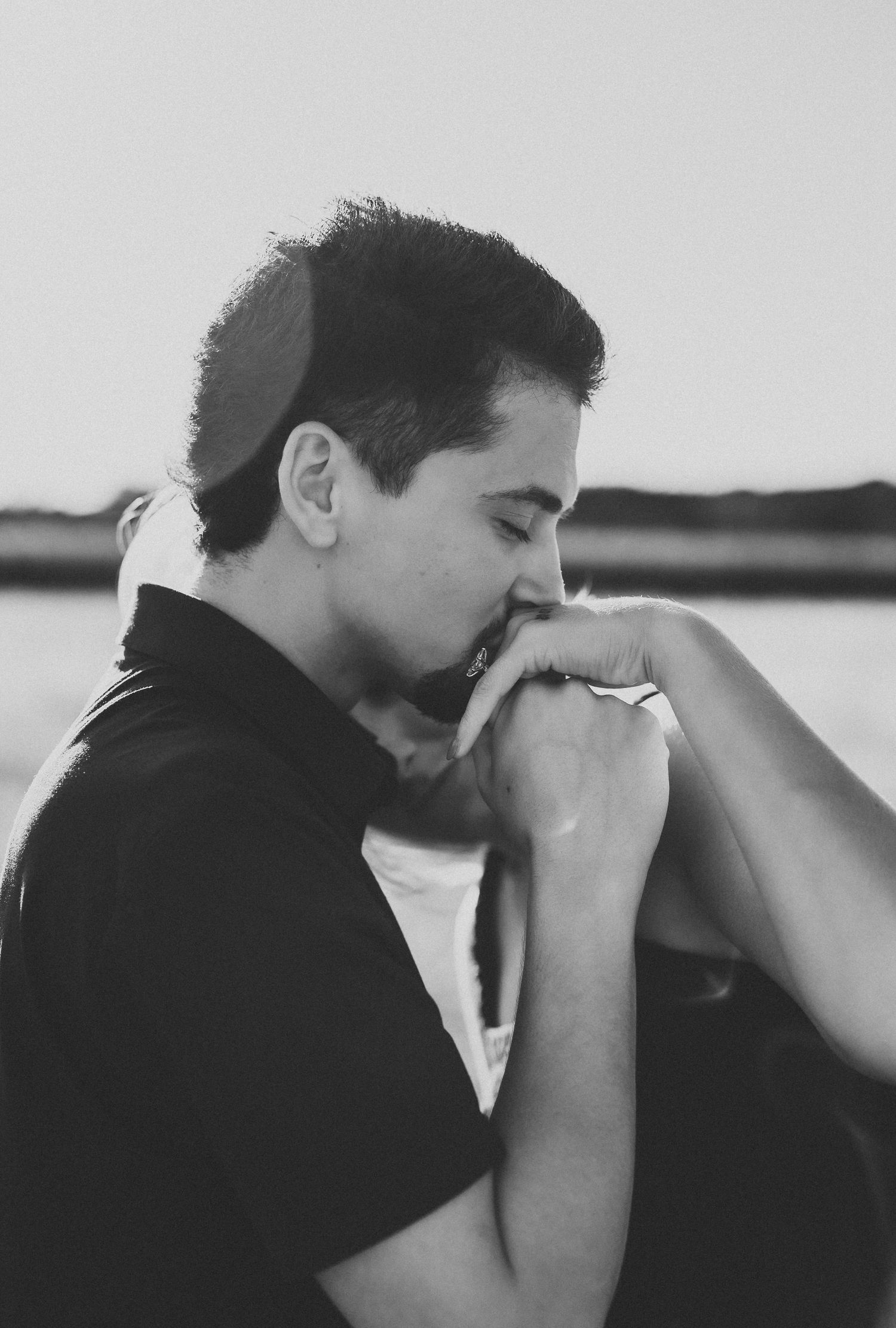 Black and white photo of a man wearing a kippah gently kissing a woman's hand by the water at sunset.