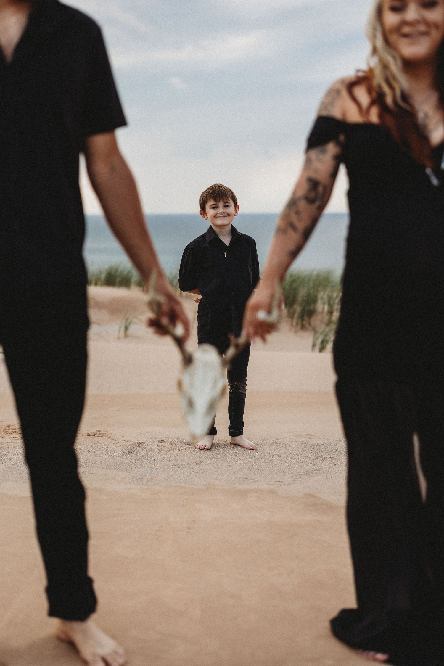 A young child in black clothing walks along a sandy beach with people in dark attire visible in the background.