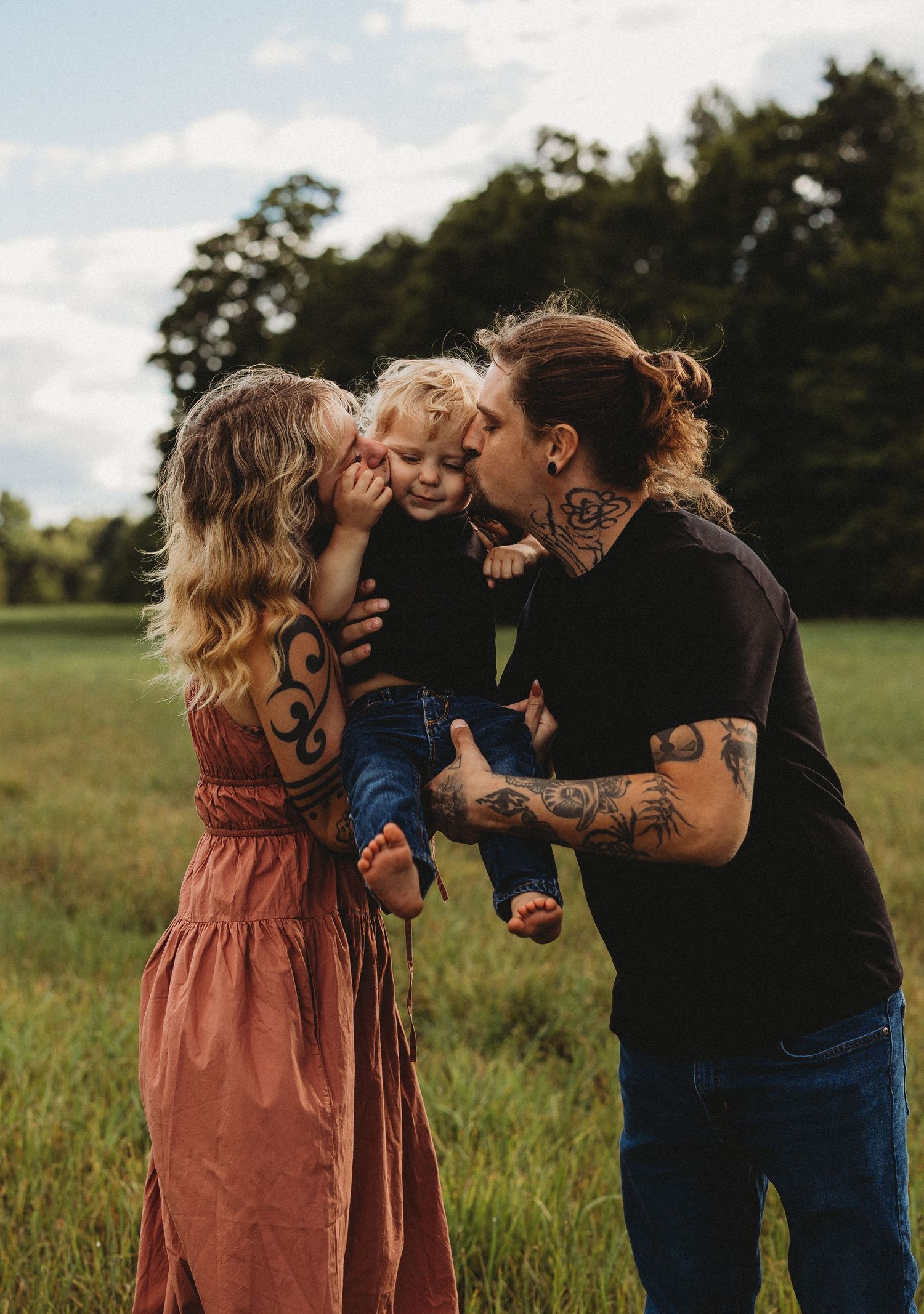 A loving family moment captured outdoors with parents kissing their small child in a rustic field setting.