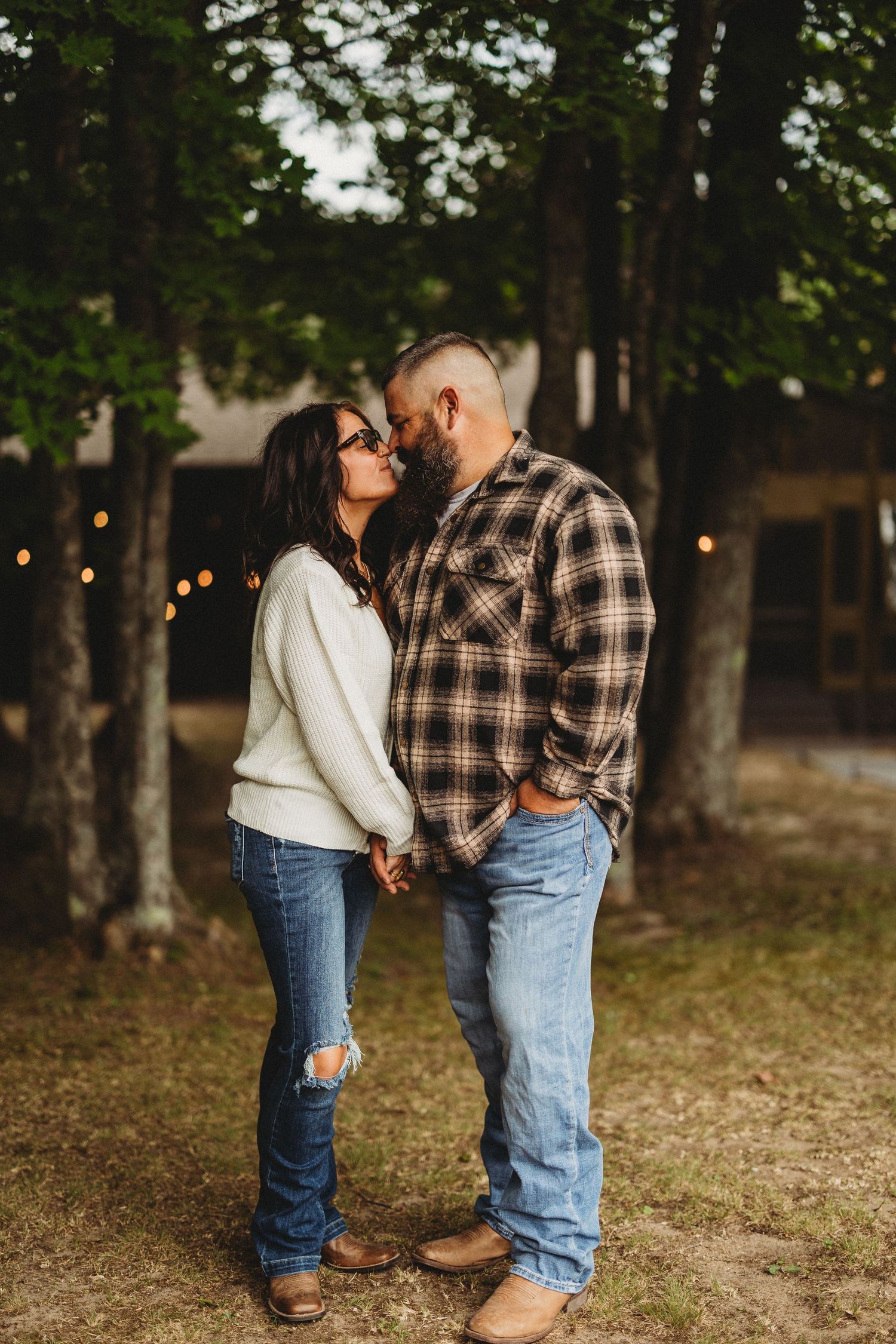 Couple in casual wear sharing intimate moments among trees with string lights.