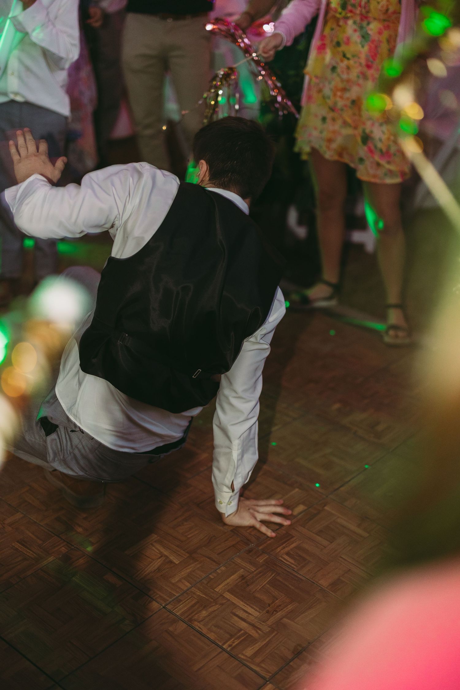 Wedding guests performing energetic dance moves on parquet floor under colorful lights.