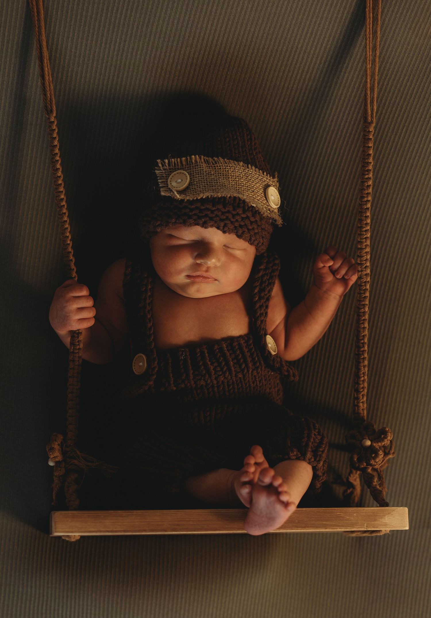 Newborn baby in brown knitted overalls and hat posed on a miniature wooden swing.