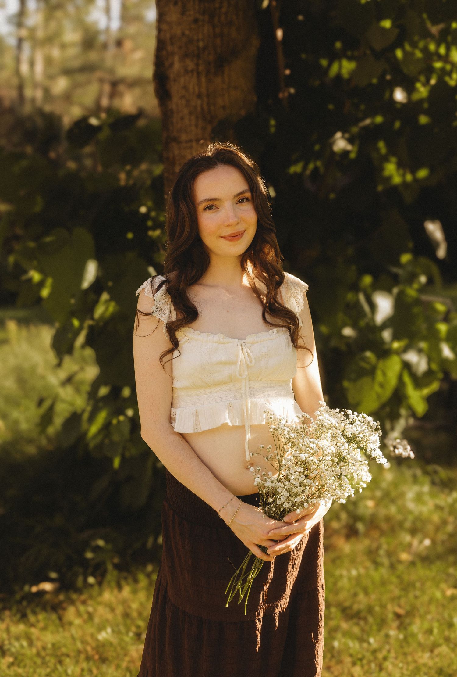 Pregnant woman in white crop top and brown skirt holding white wildflowers bouquet in sunlit garden.