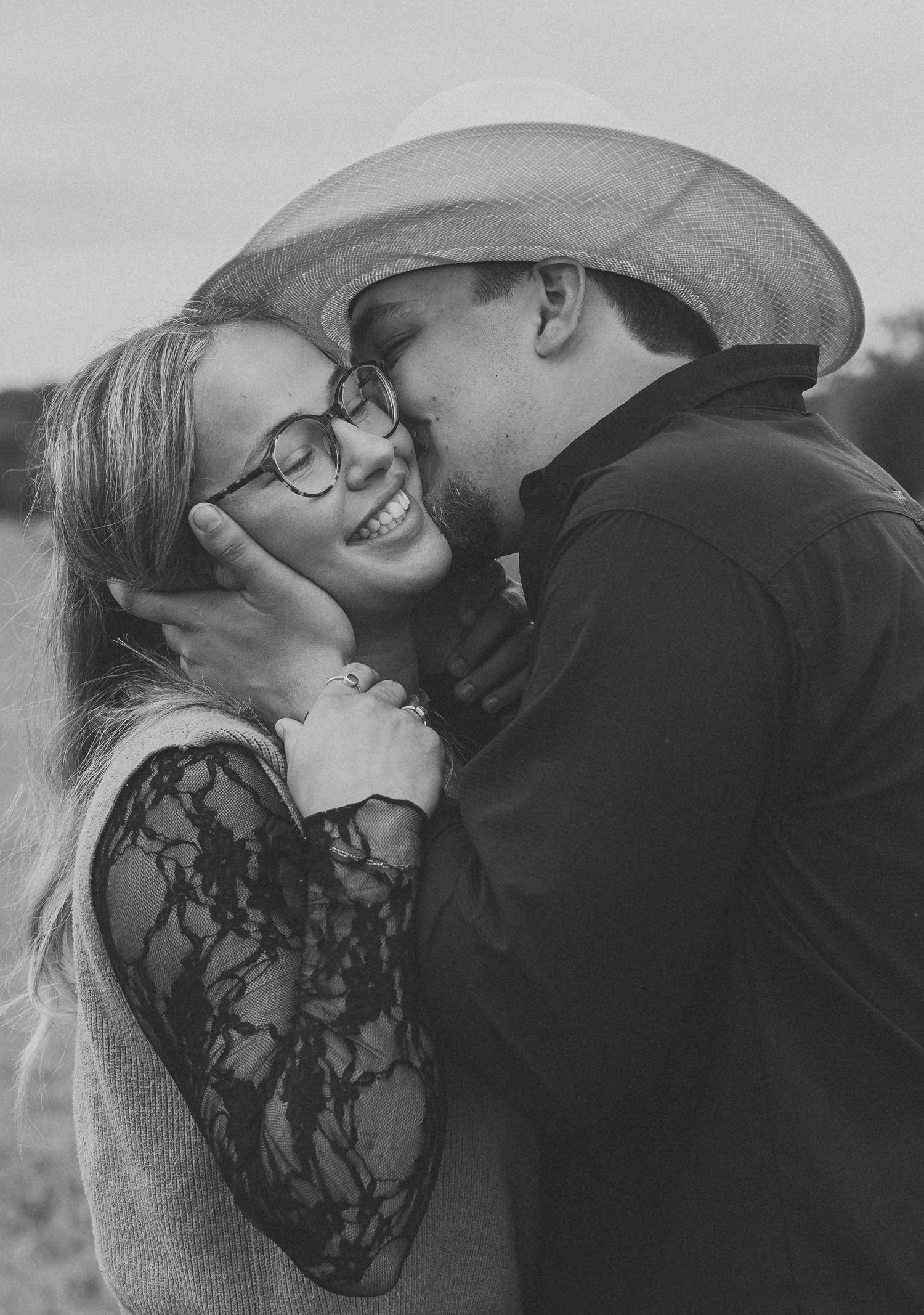 Black and white photo of a smiling woman with glasses being kissed on the cheek by a man in a cowboy hat.