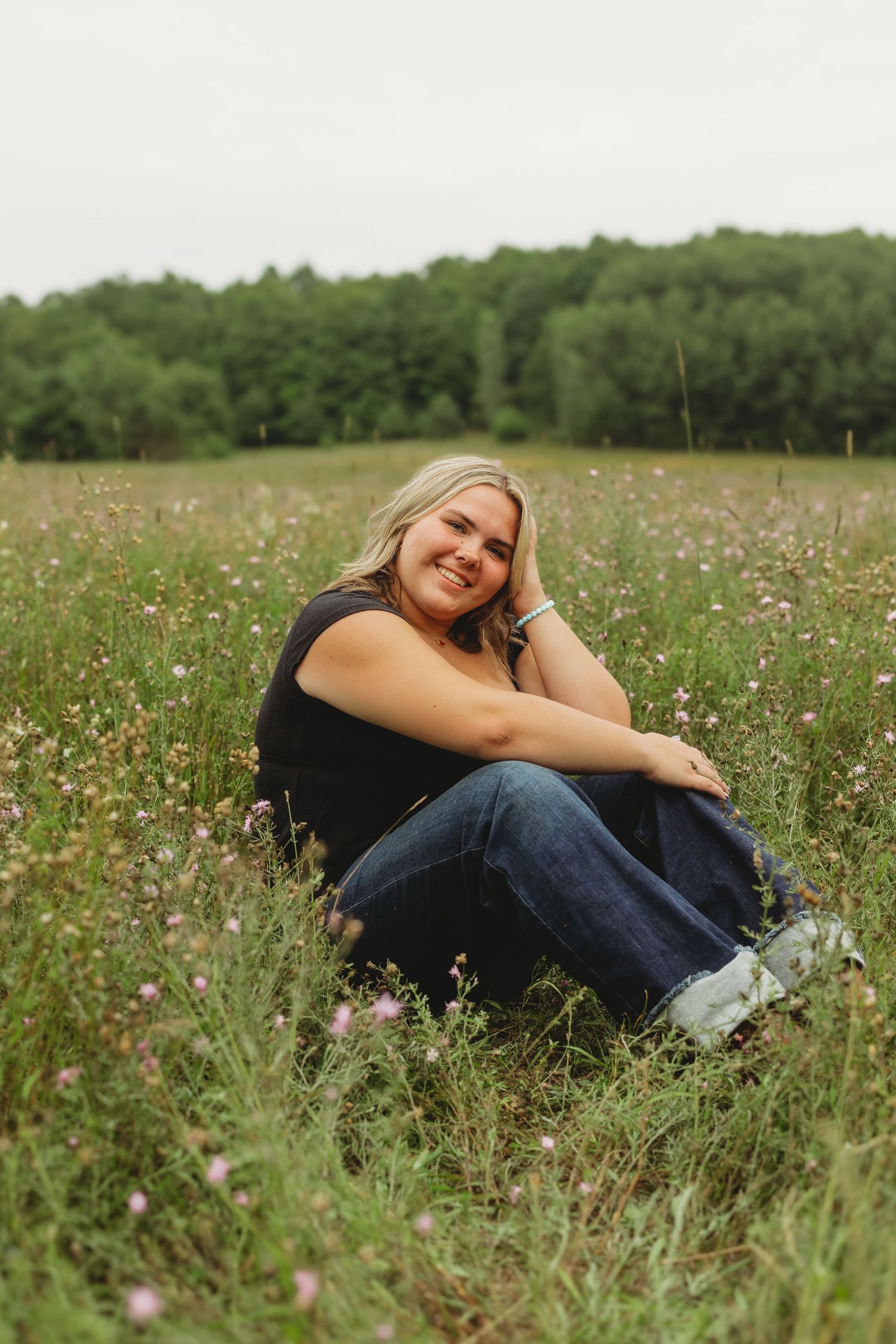 Smiling blonde woman sitting in a wildflower meadow wearing jeans and black top, with green trees in background.