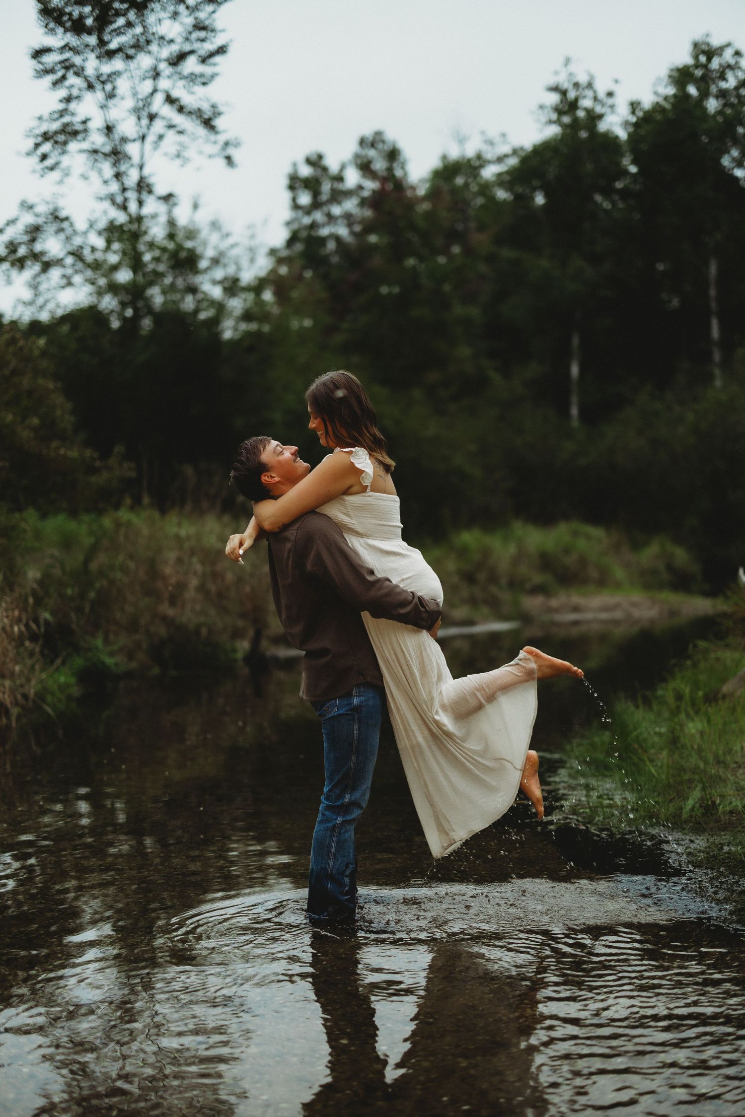 Man in jeans lifts a barefoot woman in a flowing white dress while standing in shallow water, surrounded by green trees at dusk.