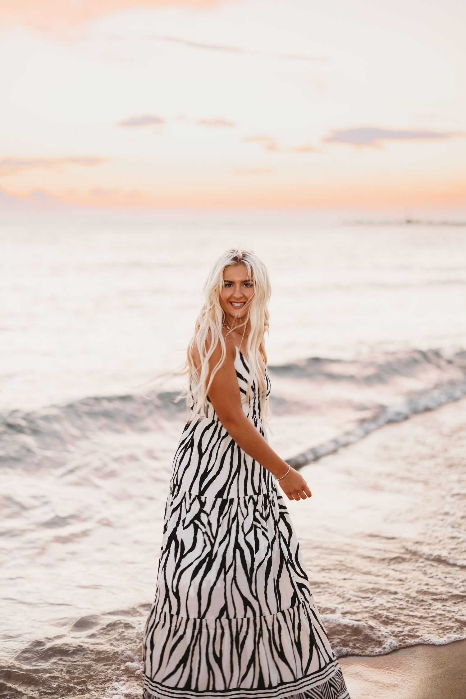 Laughing woman with long blonde hair in a zebra print dress stands in shallow ocean waves at sunset.