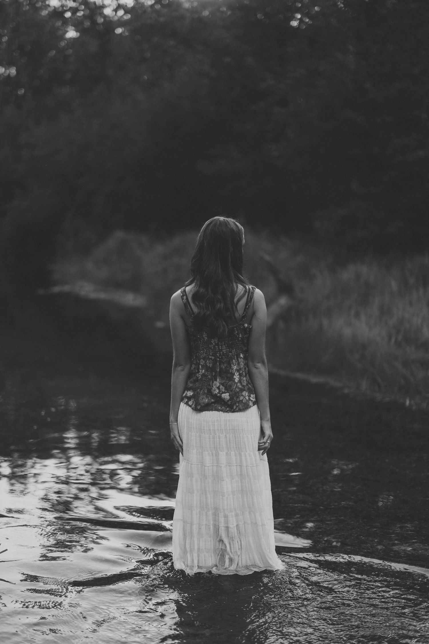 Black and white photo of a woman in a floral top and white tiered skirt standing in shallow water, seen from behind.