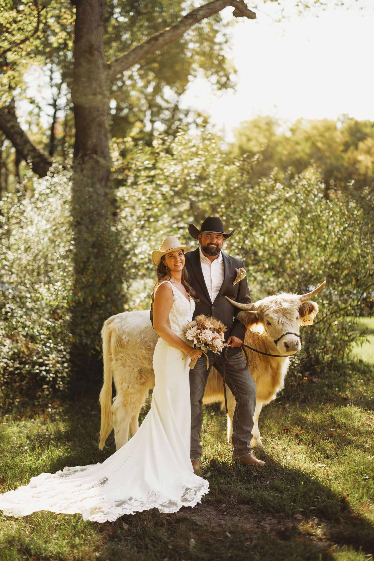 Bride in a lace wedding gown and groom in a cowboy hat pose with a shaggy highland cow among trees at a rustic outdoor wedding.