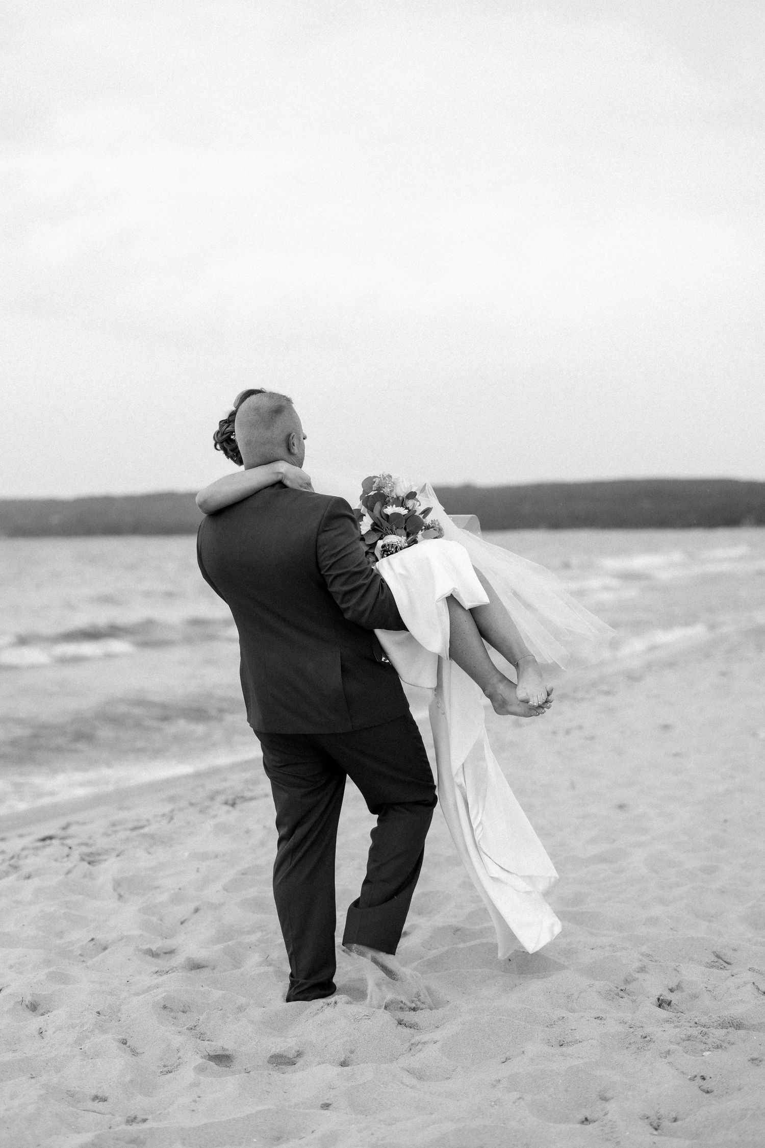 Groom carries bride in wedding dress on sandy beach in romantic black and white photo.