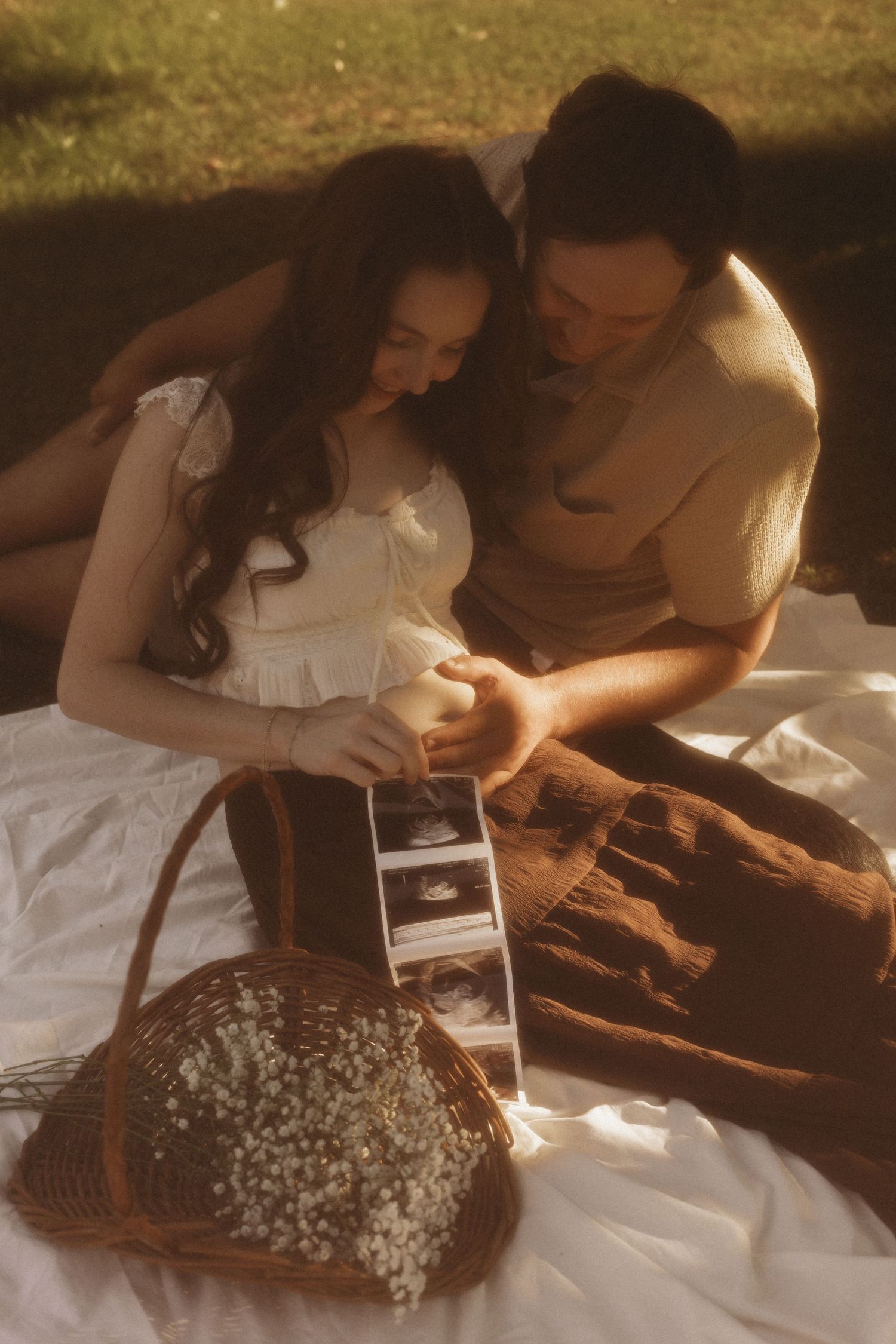 Couple sitting on white blanket outdoors, looking at ultrasound photos with a basket of baby's breath flowers nearby.
