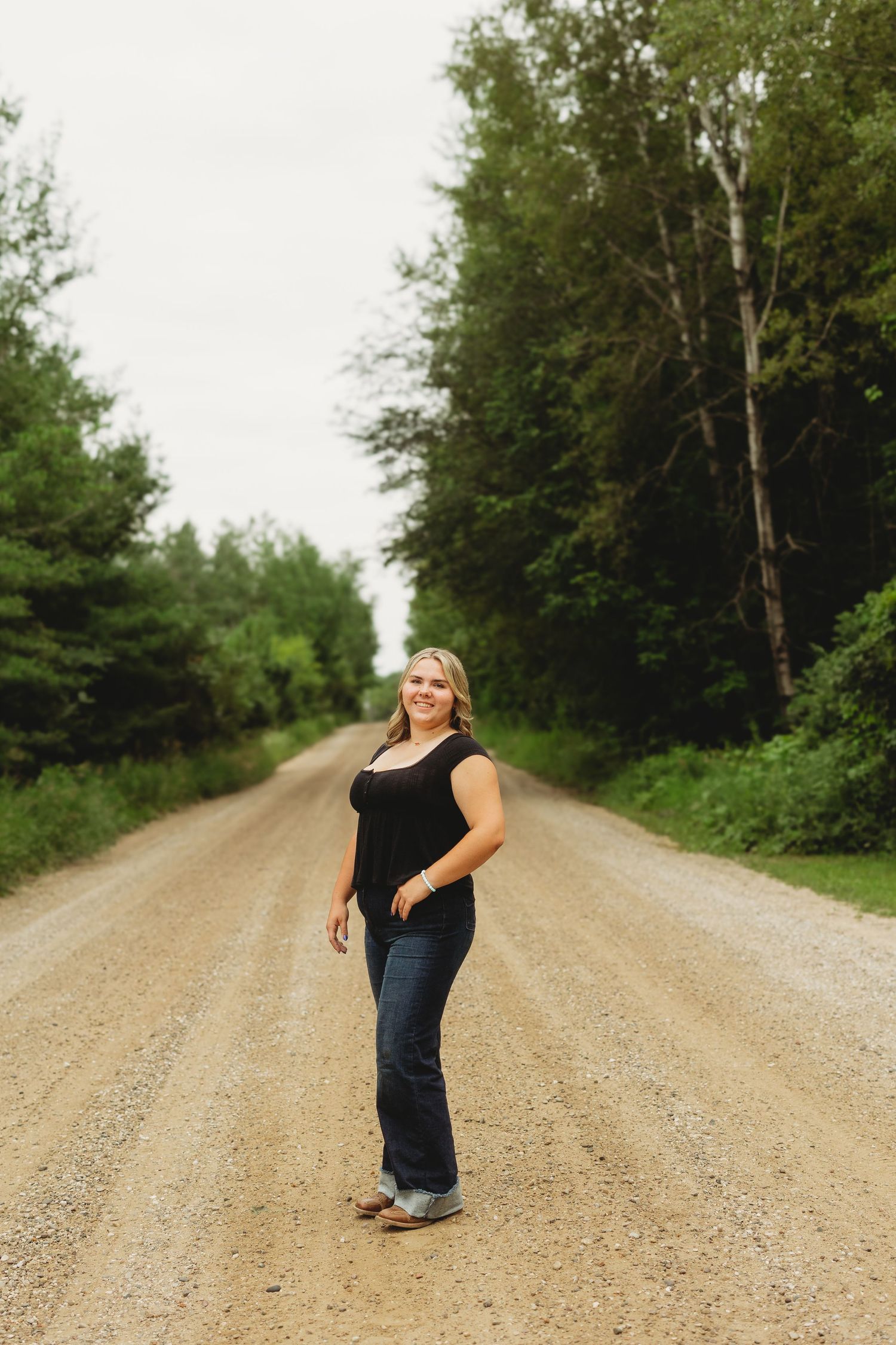 Young woman in black top and jeans posing on a rural dirt road surrounded by lush green trees.