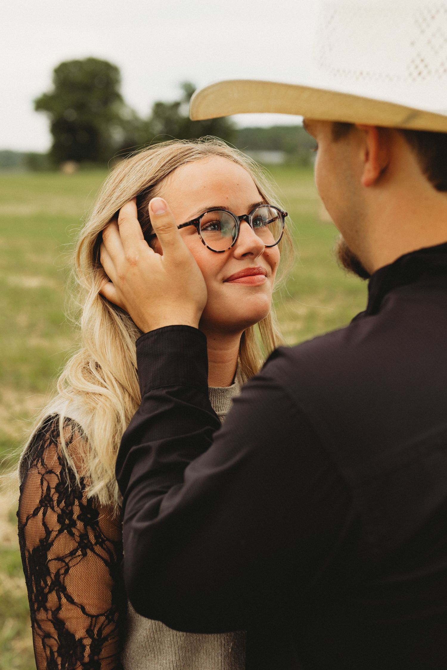 A man in a cowboy hat tenderly brushes hair from a smiling blonde woman wearing glasses in a green field.