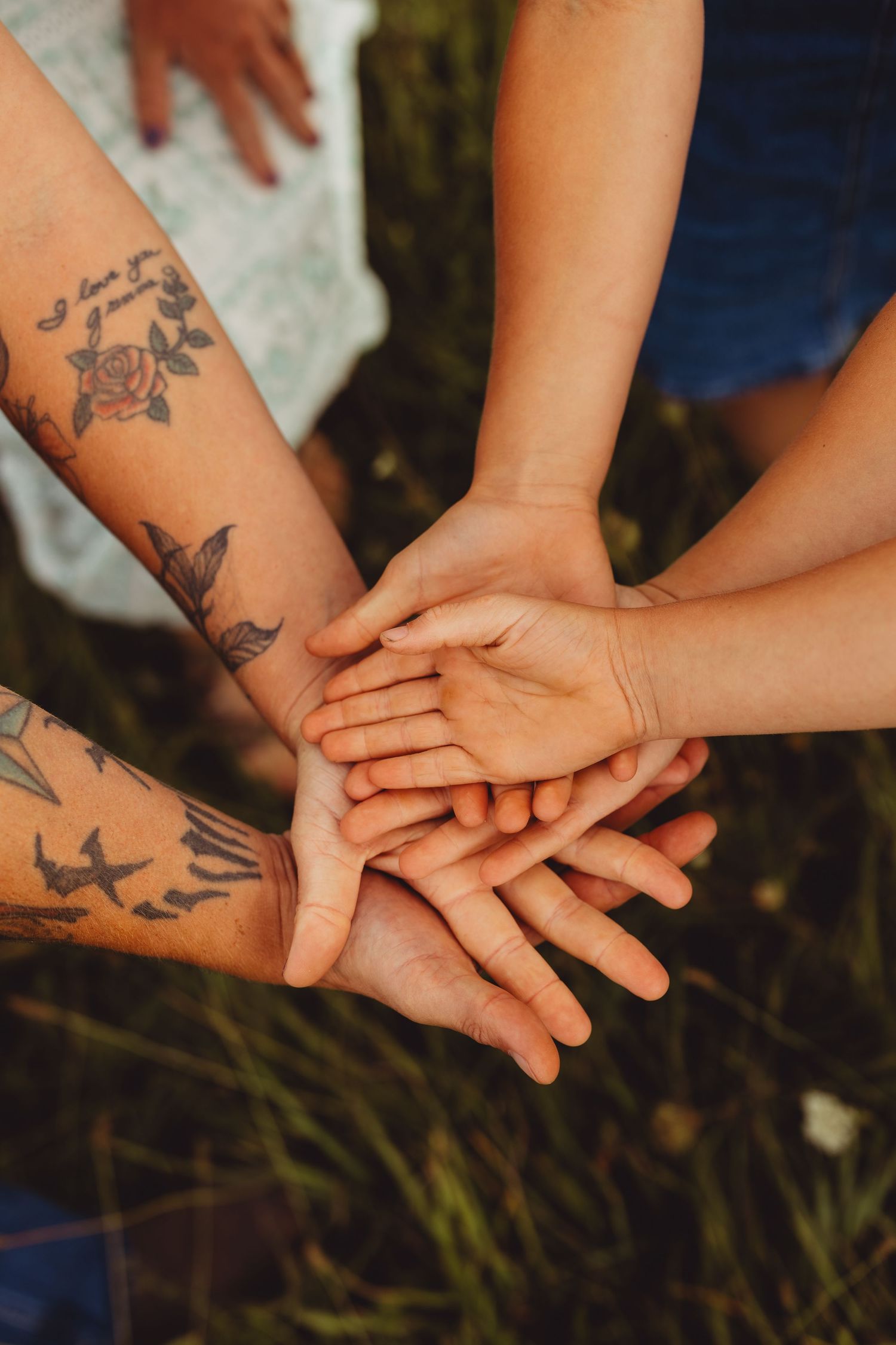 Several stacked hands, including tattooed adult arms and a child's small hands, come together in a circle over a grassy outdoor field.