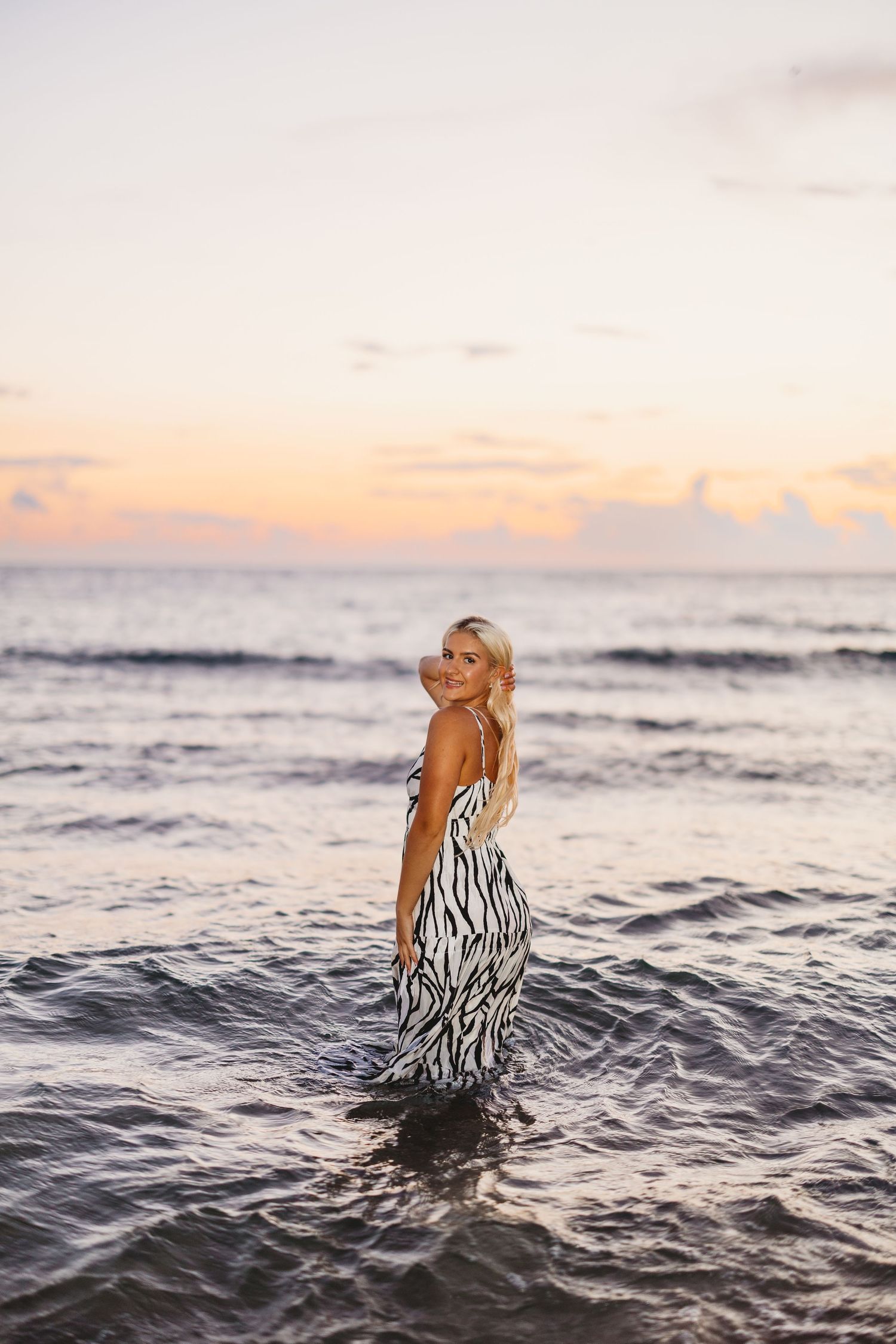 Blonde woman in a zebra print dress stands in shallow ocean water, looking over her shoulder at a pastel sunset sky.