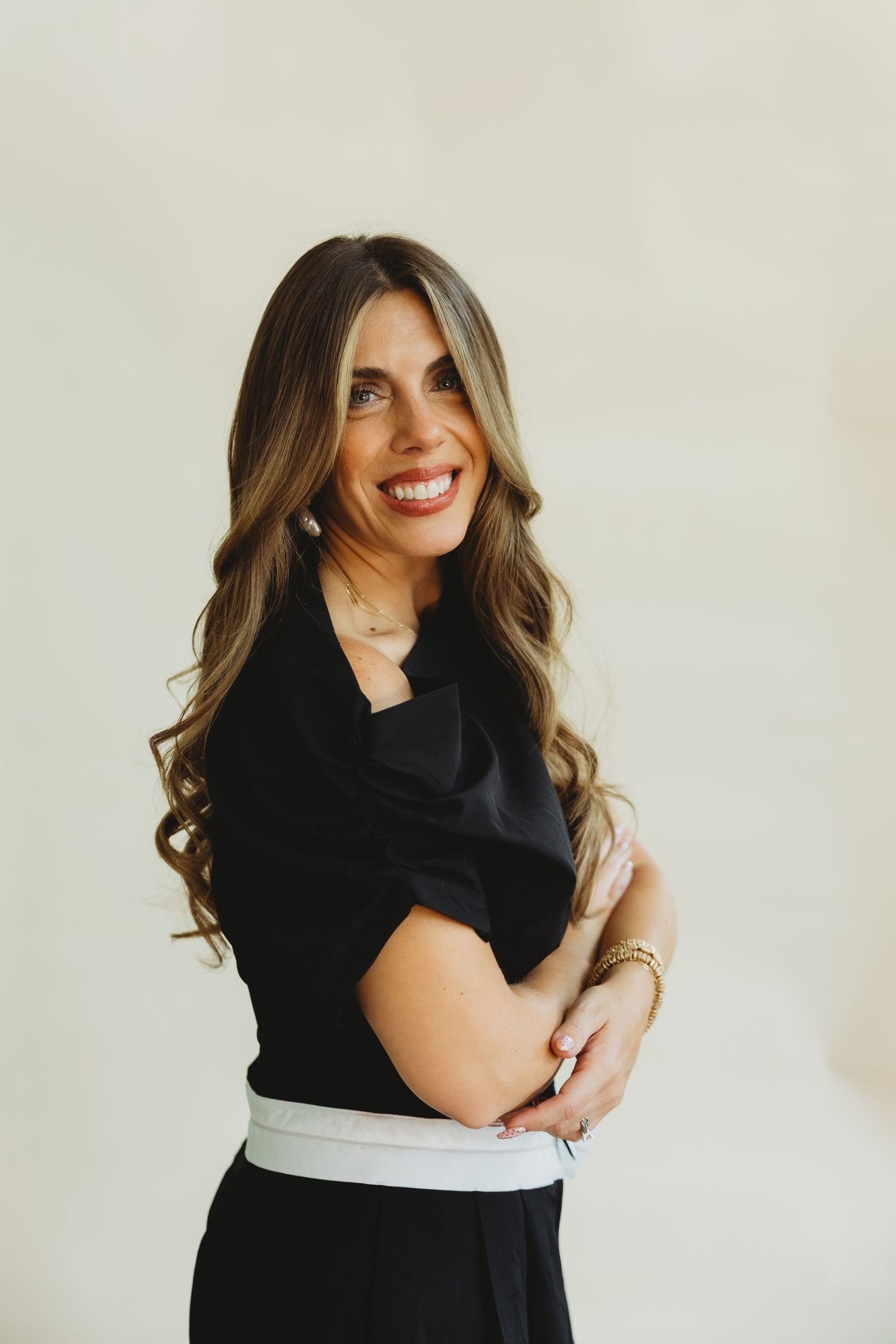 Smiling woman with long wavy hair in a black top and white-banded skirt poses with arms crossed against a neutral studio backdrop.