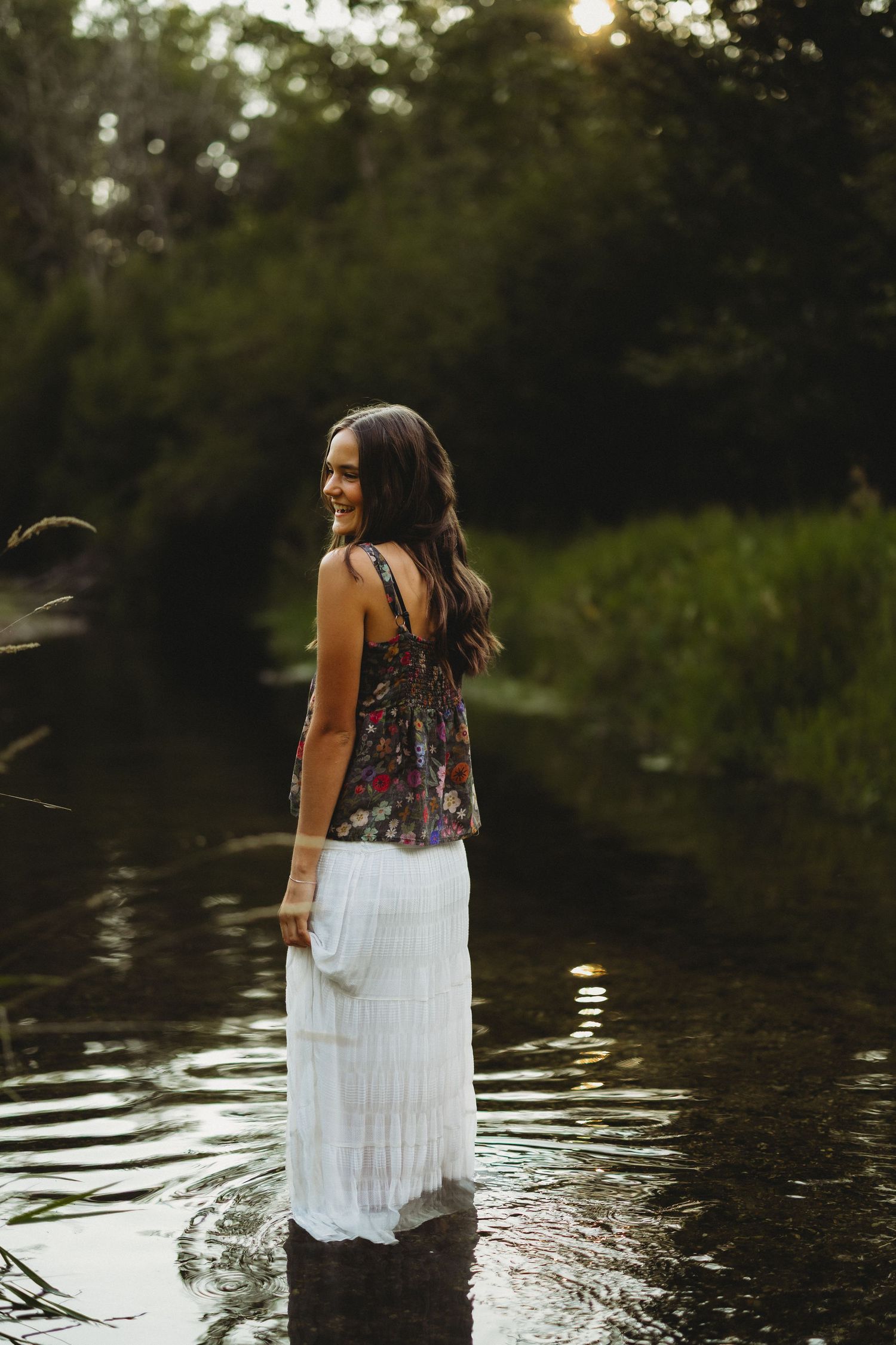 Woman in a floral top and white skirt stands in a calm woodland stream, smiling over her shoulder at sunset.