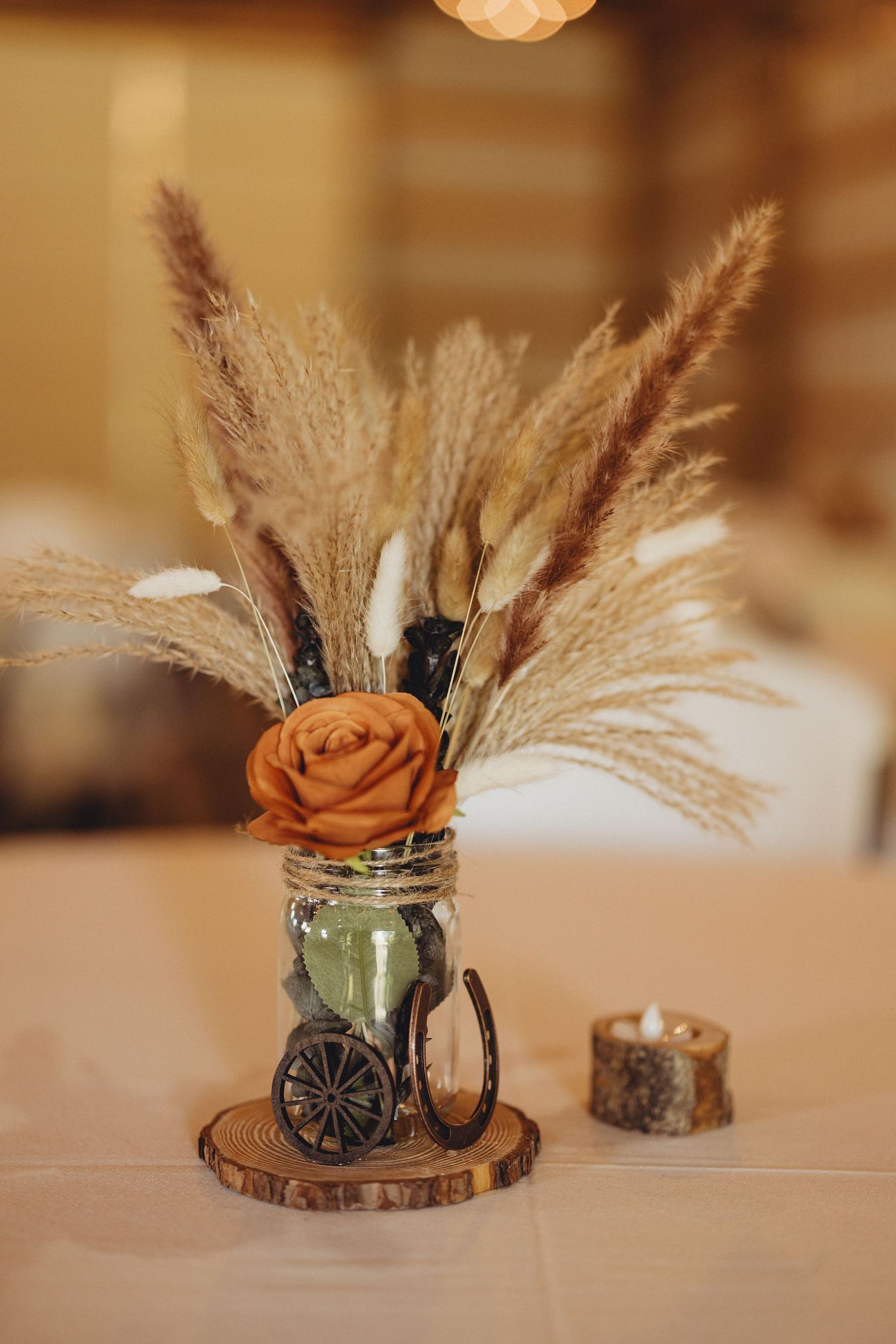Rustic wedding centerpiece of pampas grass and an orange rose in a twine-wrapped jar with a horseshoe, on a wood slice beside a candle.