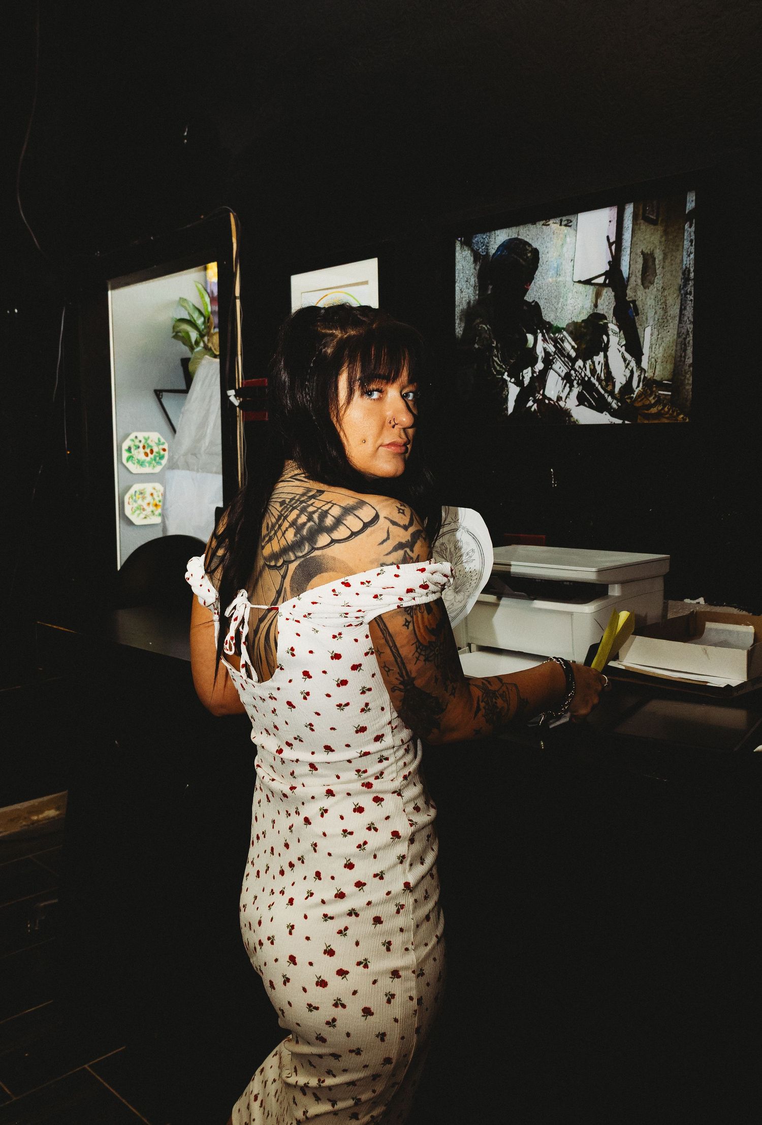 Tattooed woman in floral dress posing sideways showing back tattoos in dimly lit room with artwork on wall.