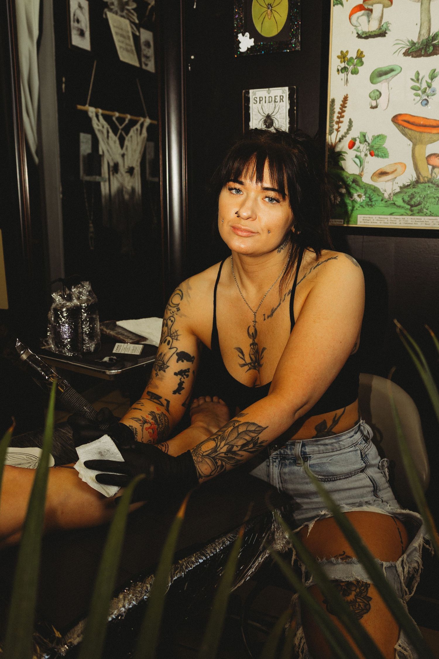 Tattooed woman in black crop top and denim shorts sitting in a tattoo studio with mushroom art on the wall.