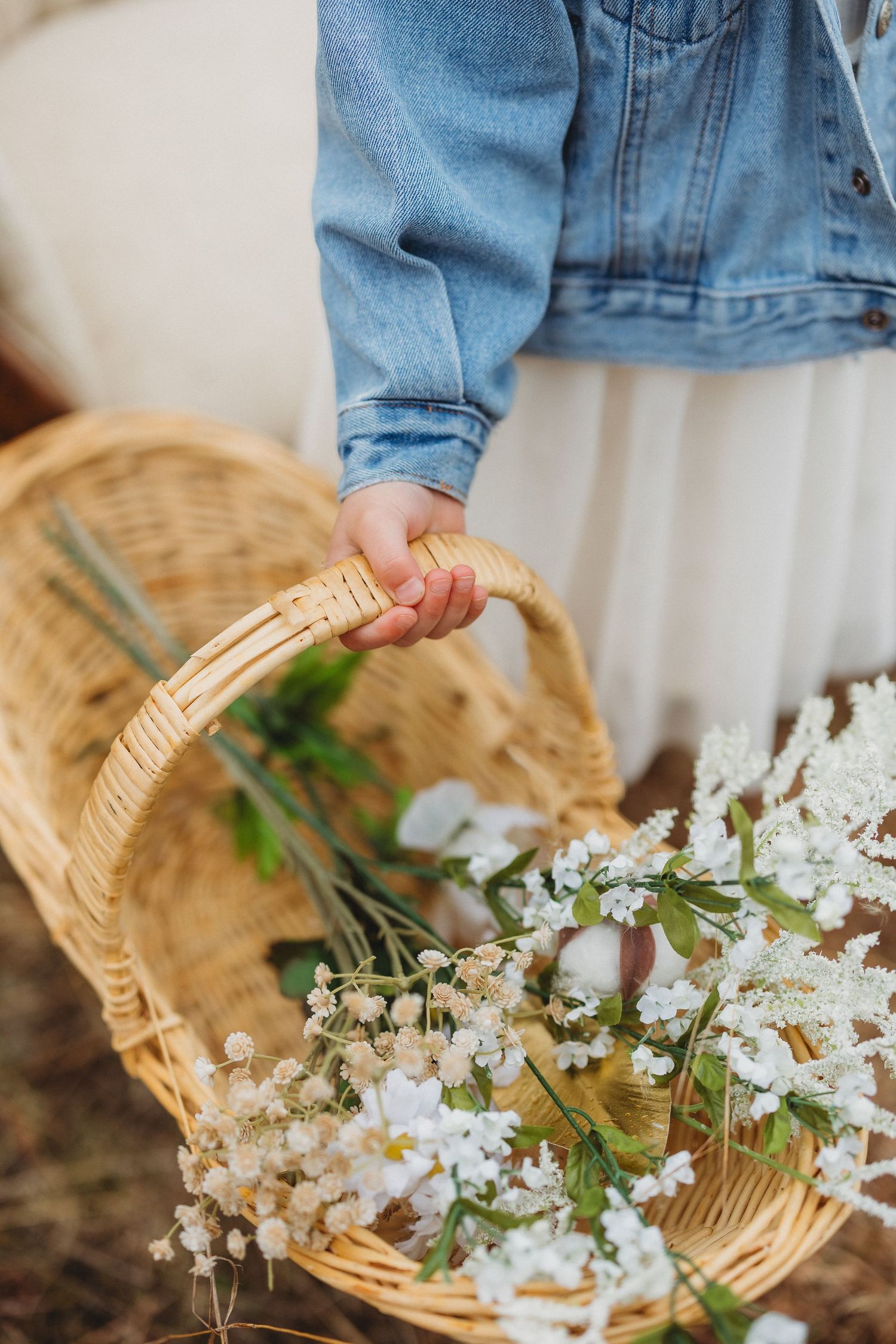 Child in denim jacket holding a wicker basket filled with white wildflowers and greenery outdoors.