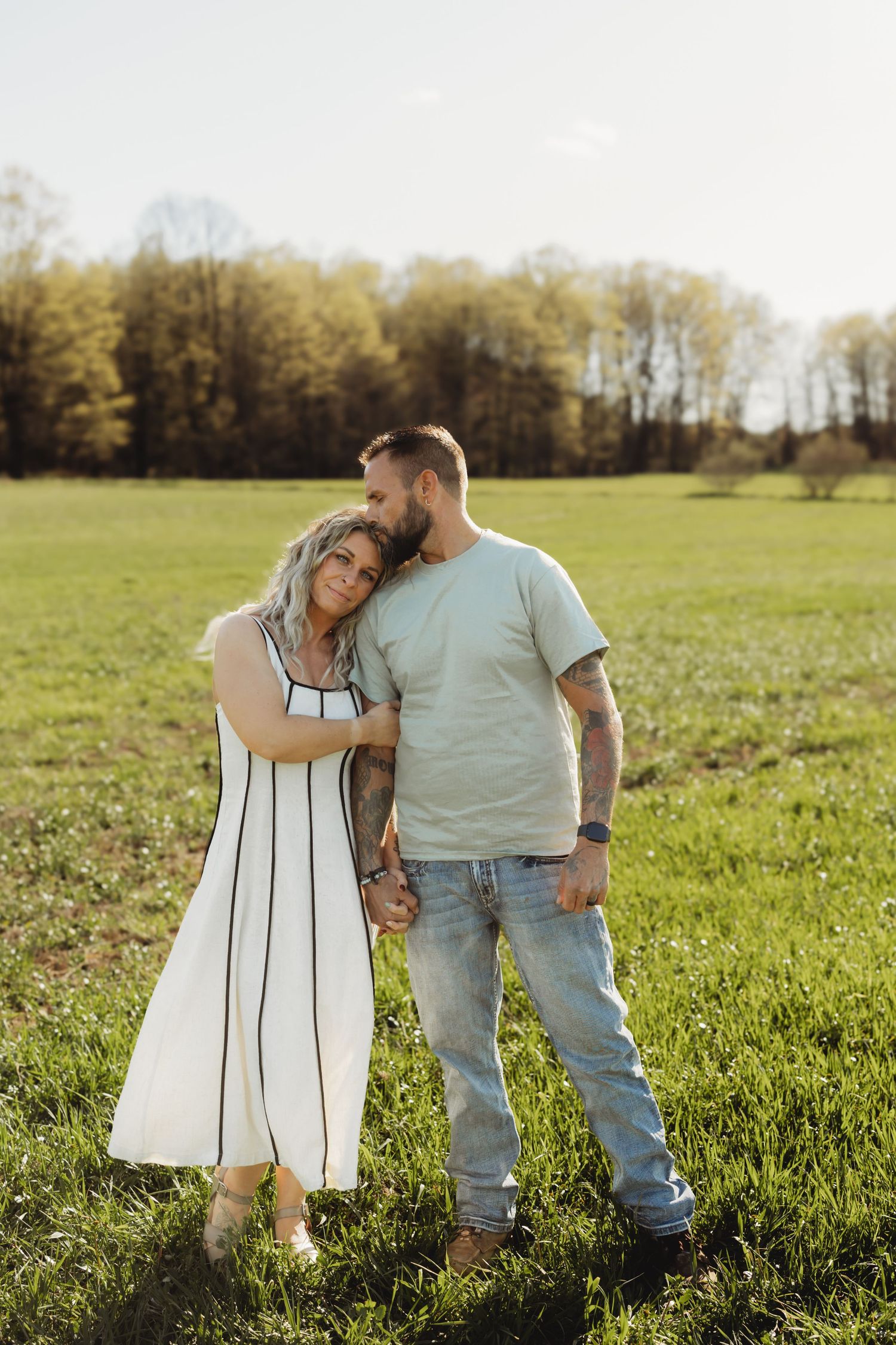 Couple embracing in a green field, woman in white dress leans on man in light tee and jeans, trees in background.
