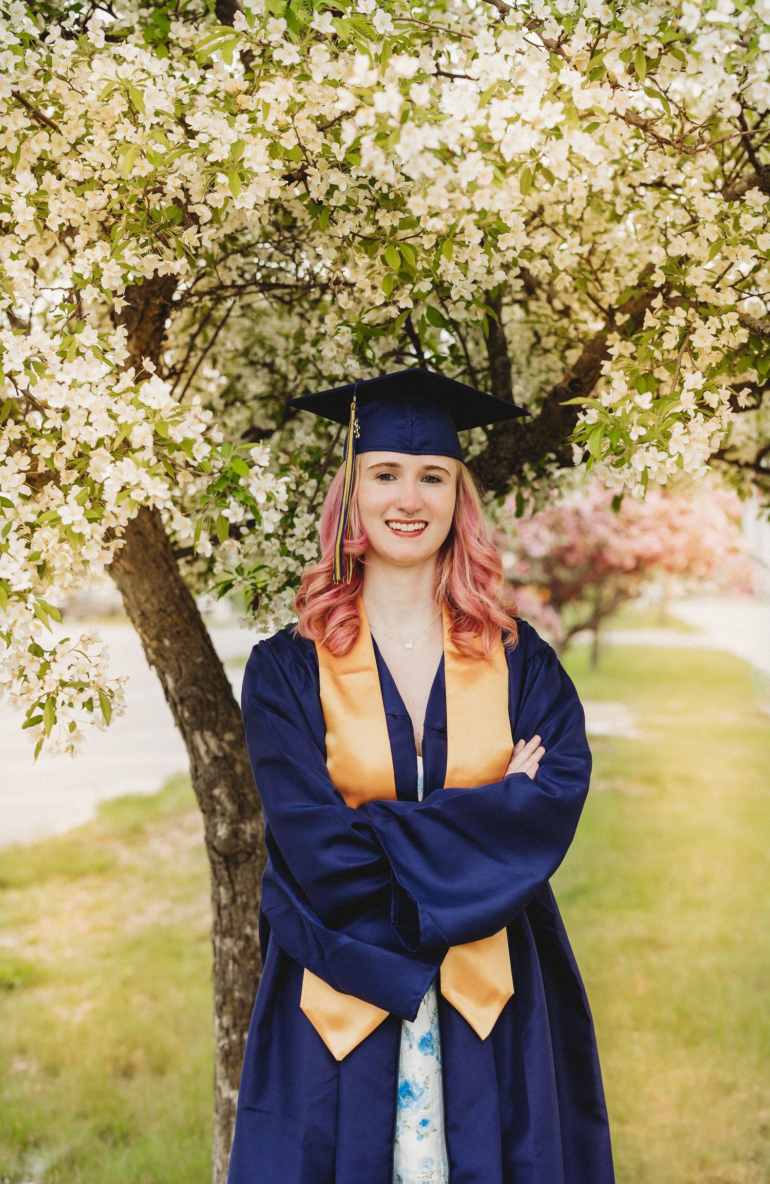 Graduate in navy blue cap and gown poses under blooming white cherry blossom trees in spring.