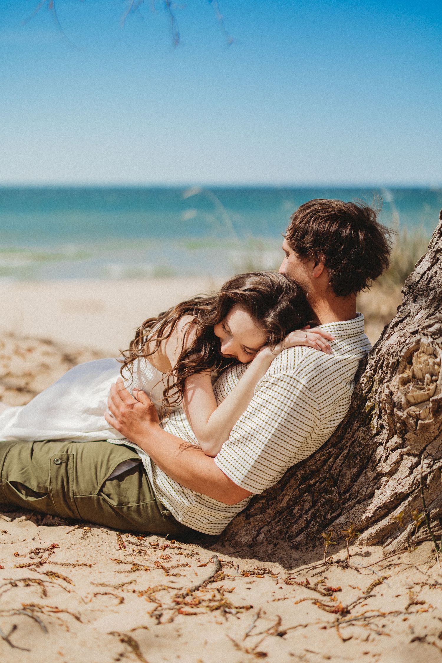 Couple resting together against driftwood on sunny beach with ocean view.