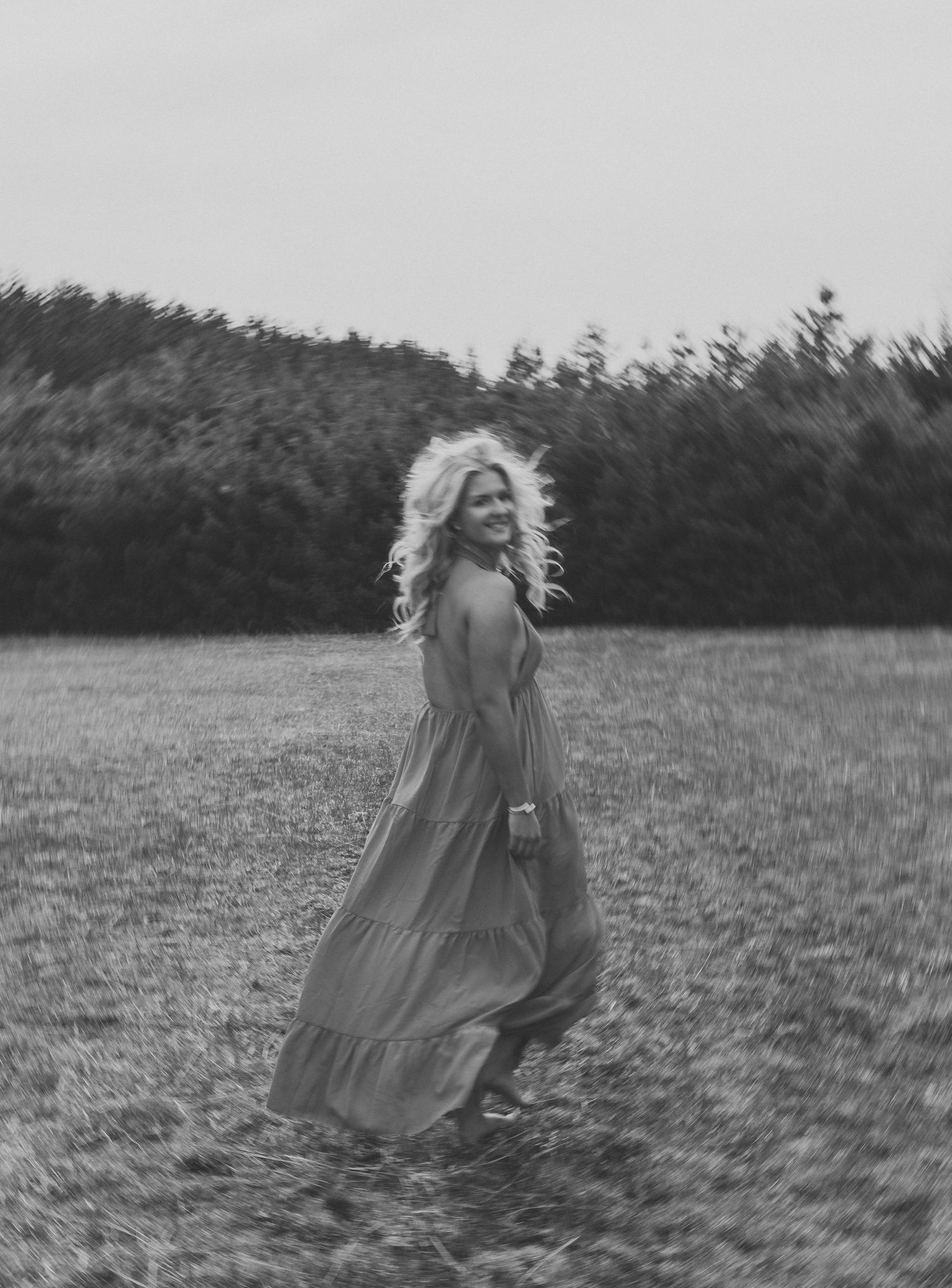 Black and white photo of a blonde woman in a flowing maxi dress twirling in a grassy field with trees behind her.