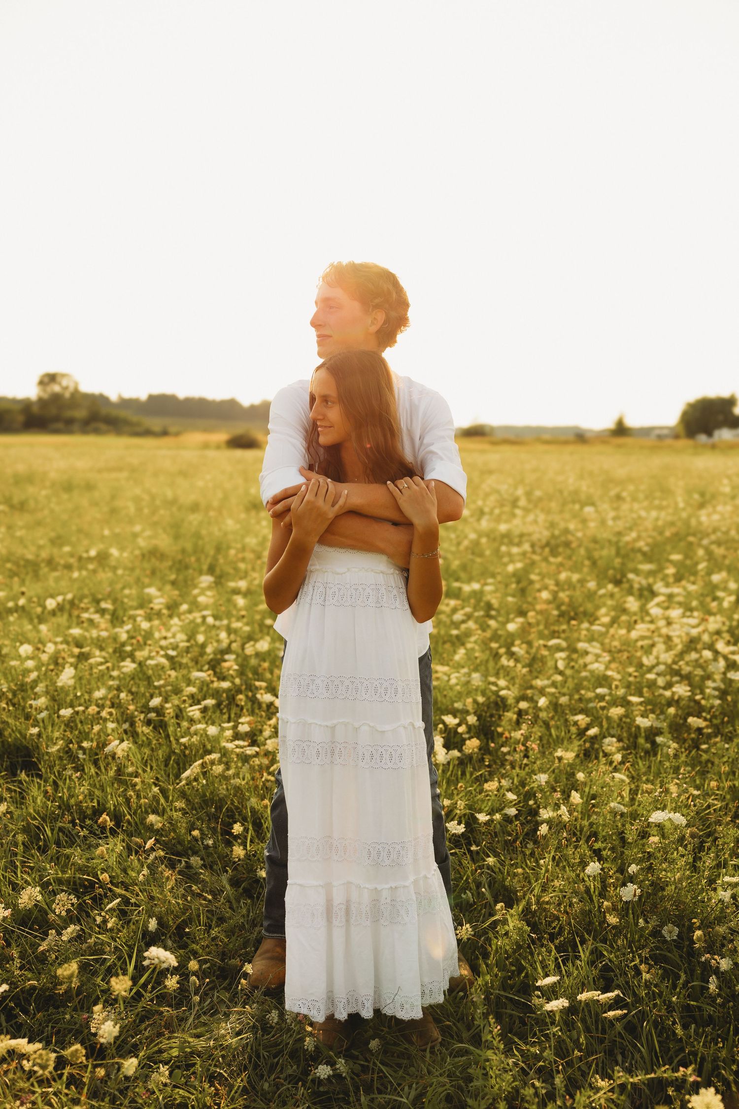 Couple embracing in a golden sunlit wildflower field, woman in white dress, man in white shirt at sunset.