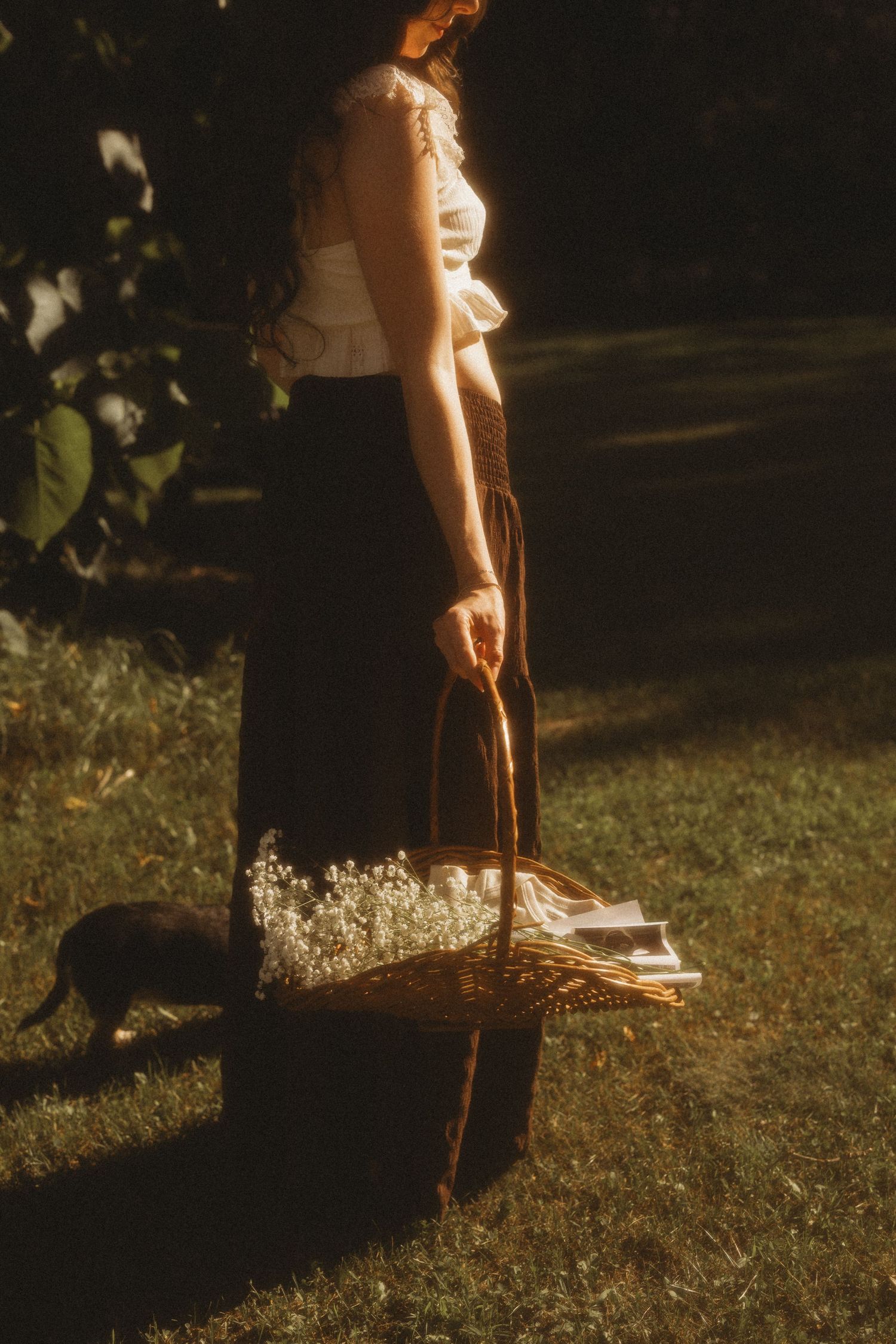 Woman in white top and brown skirt holding wicker basket with wildflowers on sunny grass lawn.