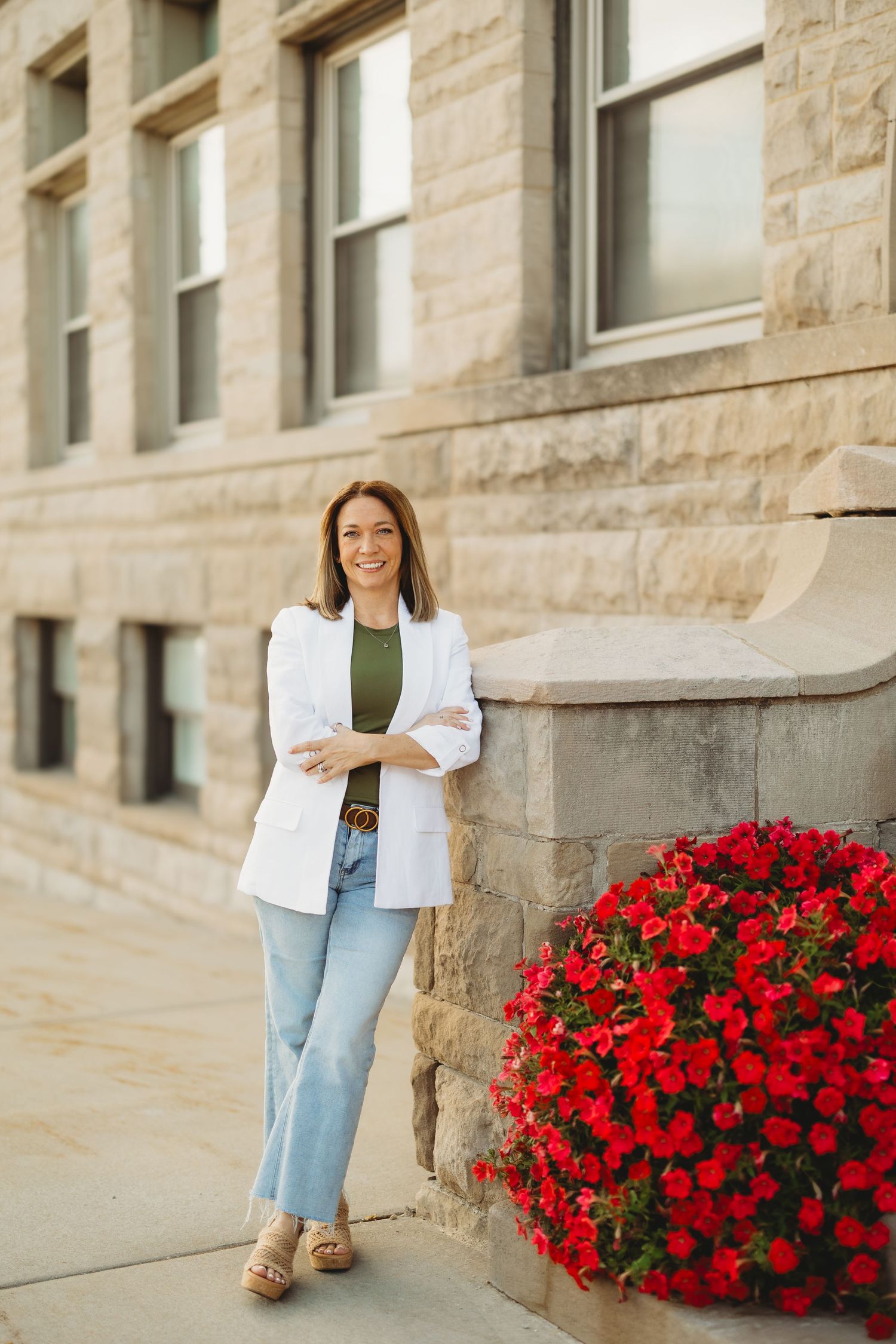 Woman in white blazer and jeans leaning against stone building with red flowers, smiling outdoors.