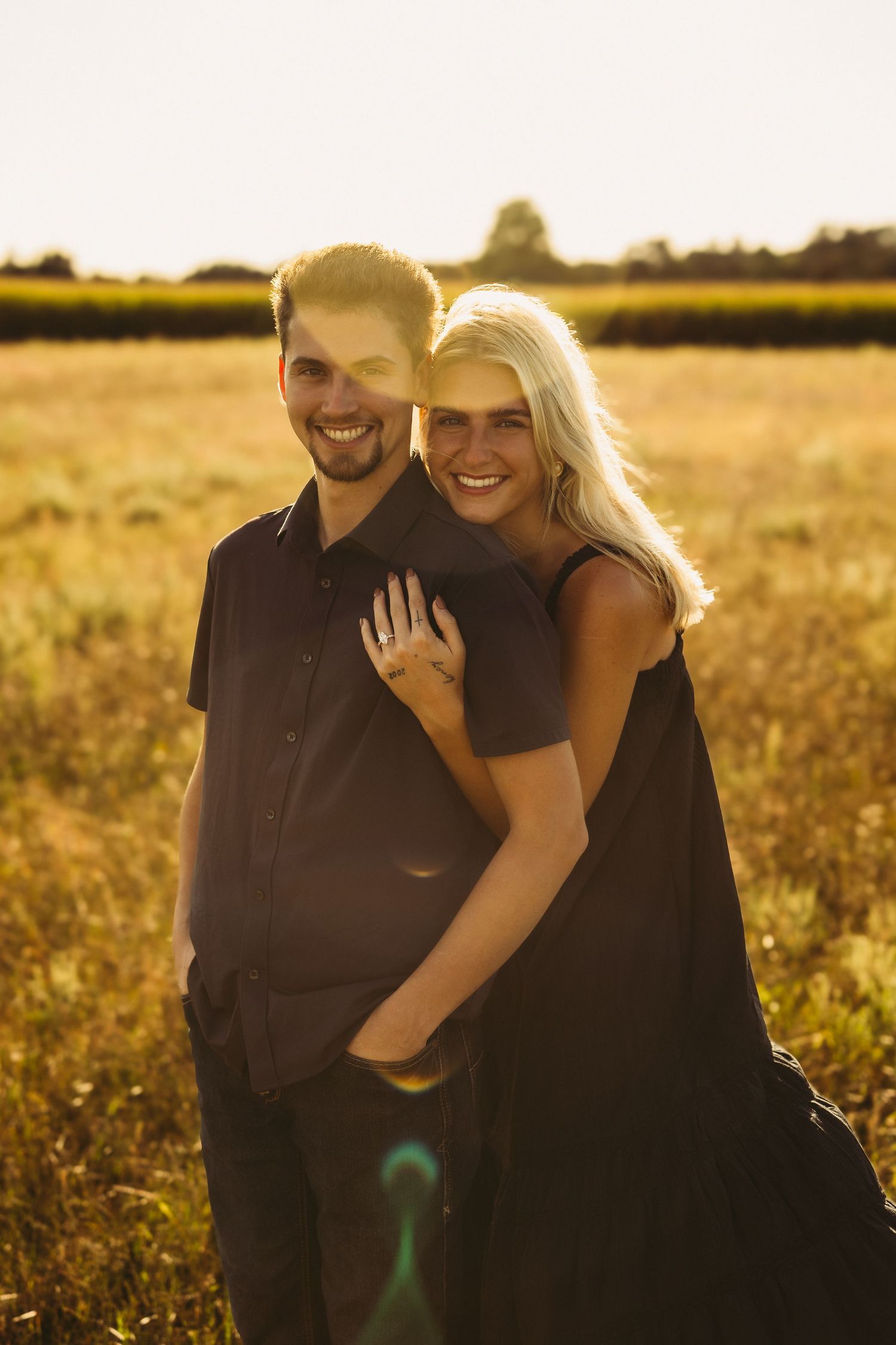 Smiling couple embracing in a golden wheat field at sunset, the woman in a black dress hugging the man from behind.