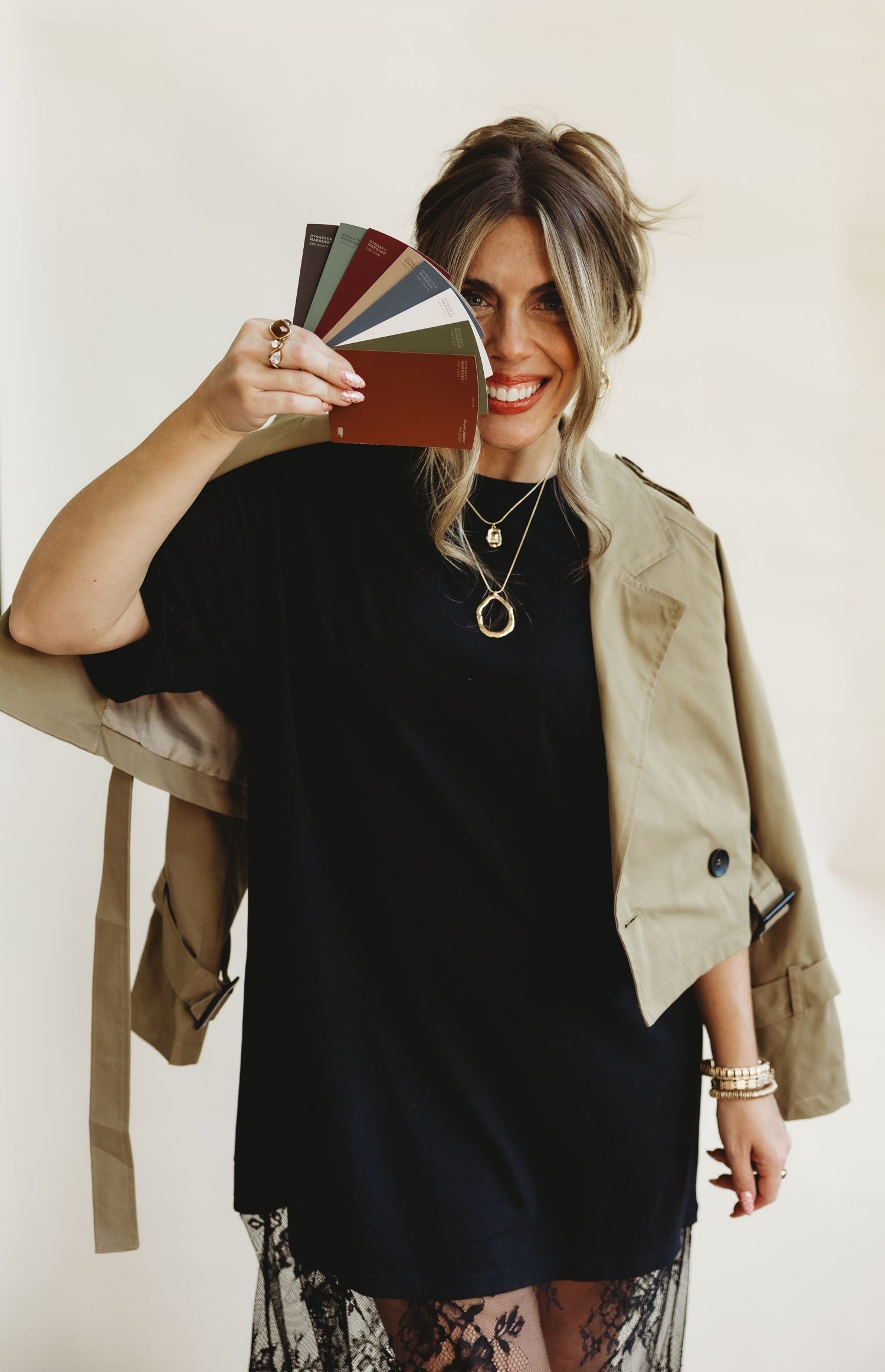 Smiling woman in a black tee dress, lace skirt and tan cropped jacket holds a fan of color swatch cards over one eye.