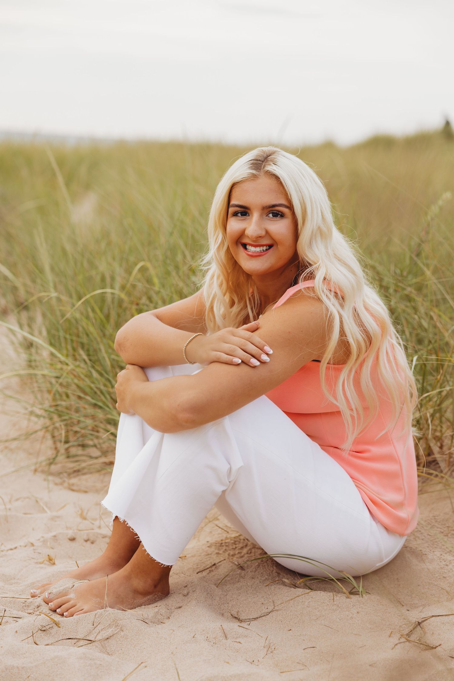Smiling woman with long blonde hair in a coral top and white pants sits barefoot on a sandy beach beside tall dune grass.