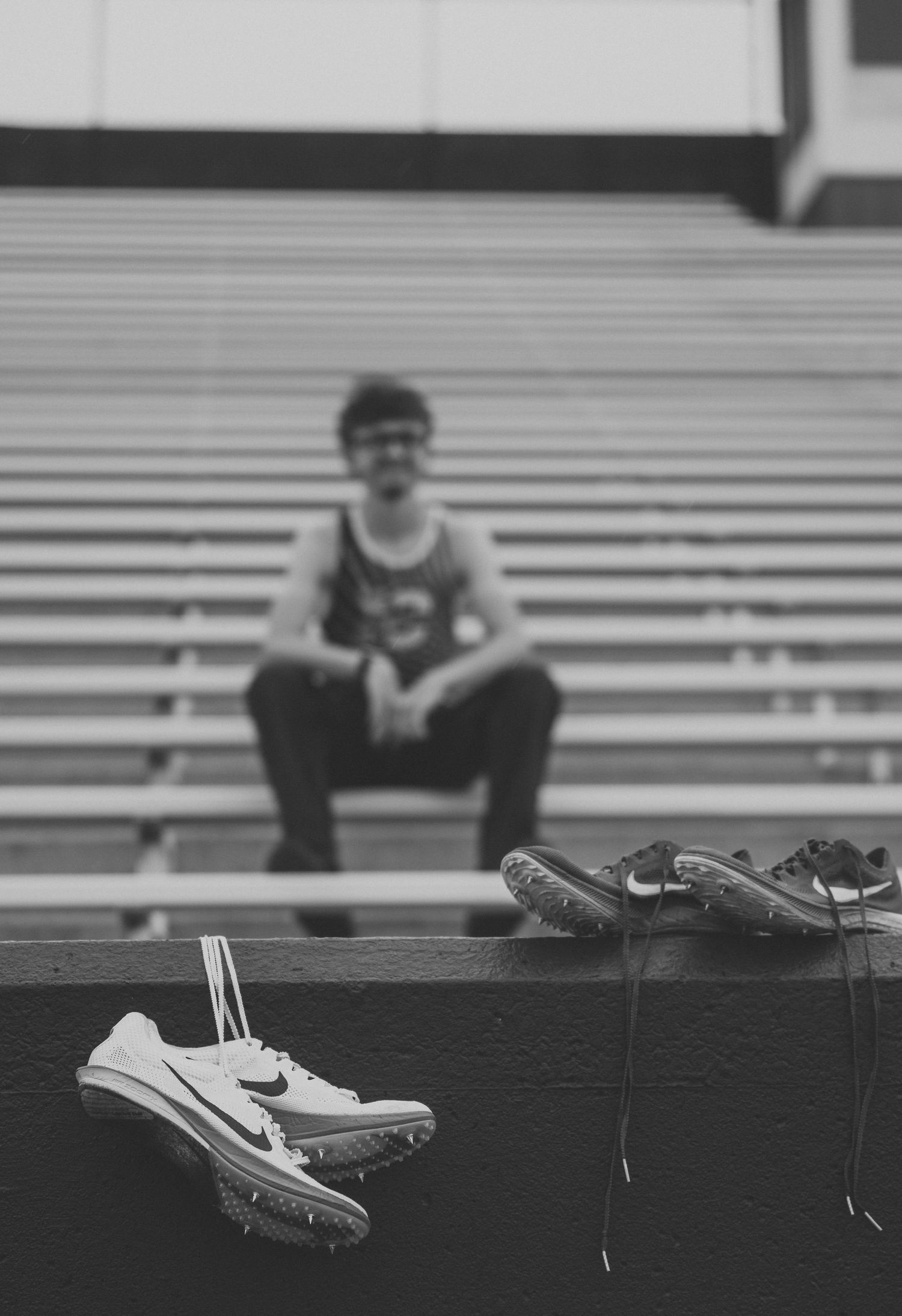 Black and white photo of a runner sitting on empty stadium bleachers behind blurred track spikes resting on a concrete ledge.