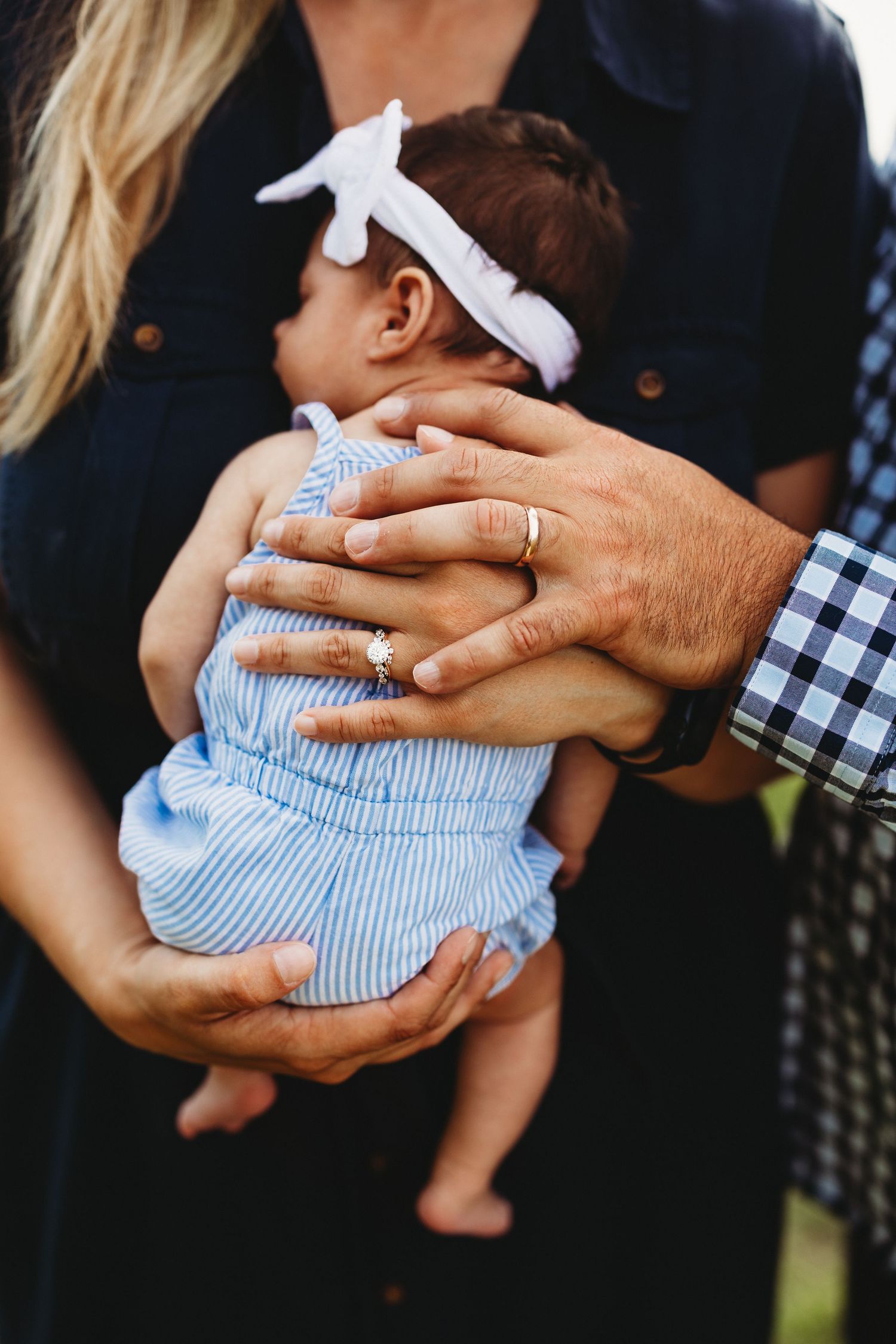 Close-up shots of parents cradling baby wearing blue dress and white bow in outdoor setting.