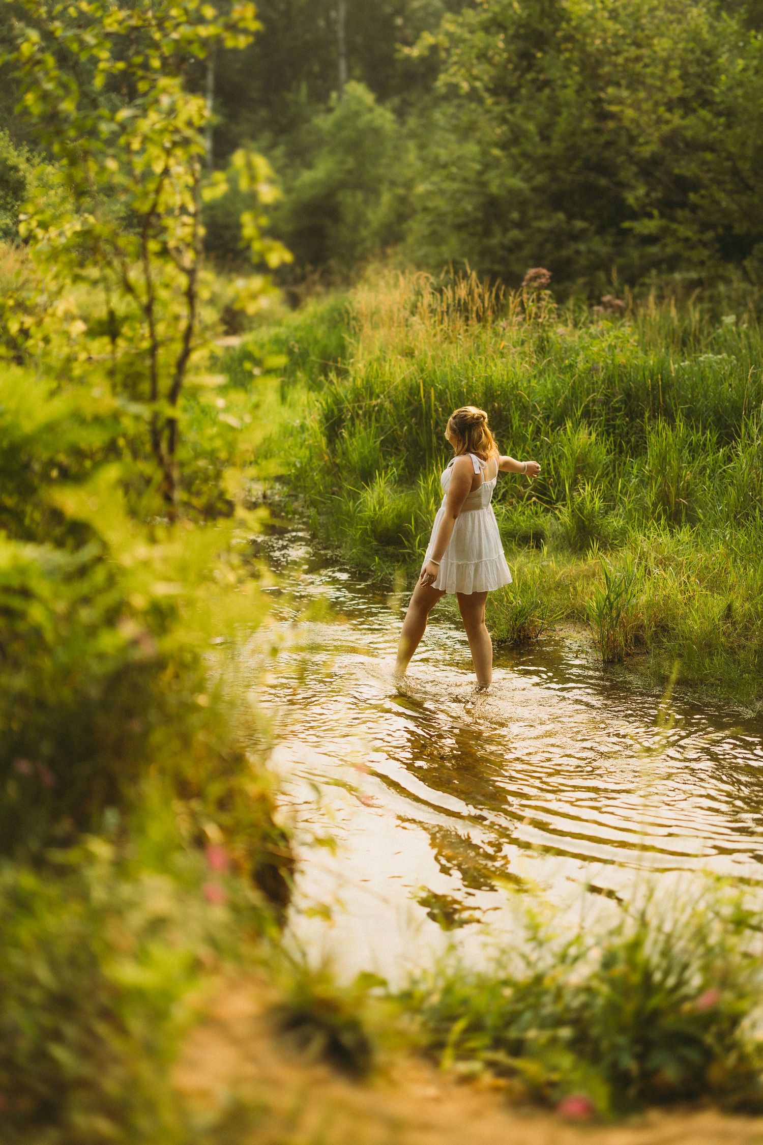 Figure in white dress wading through sunlit water surrounded by lush greenery.