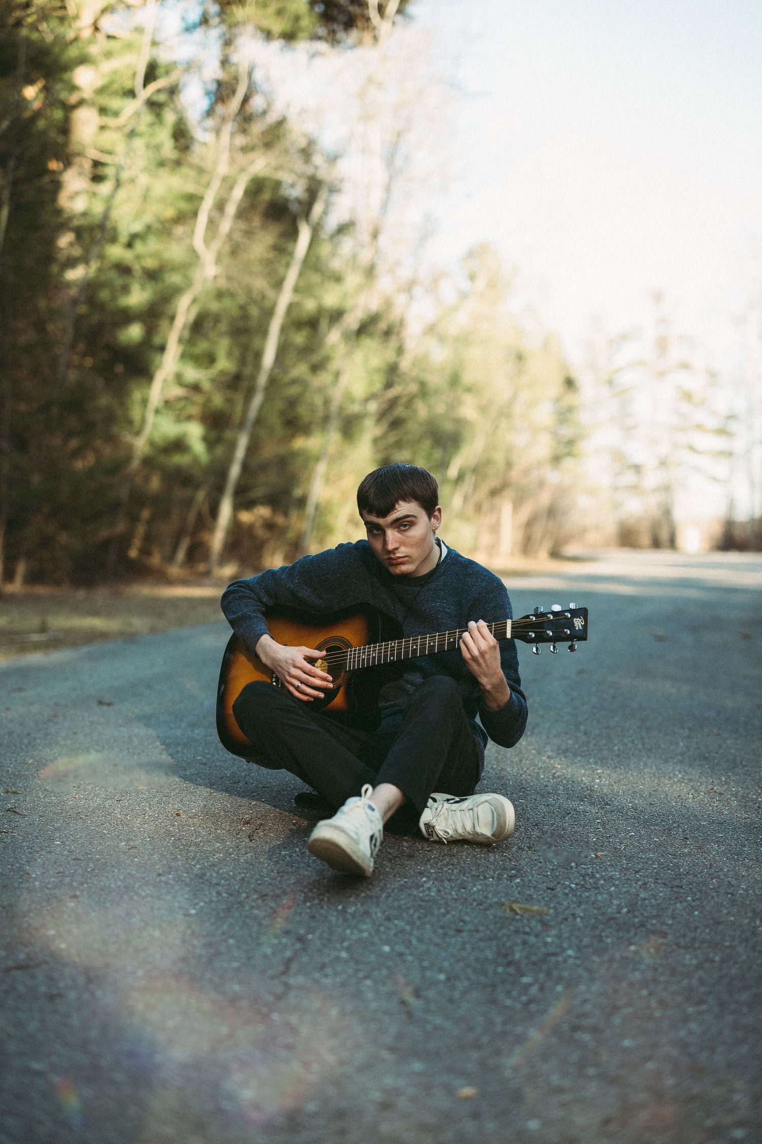 Musician playing acoustic guitar while sitting on asphalt road surrounded by trees.
