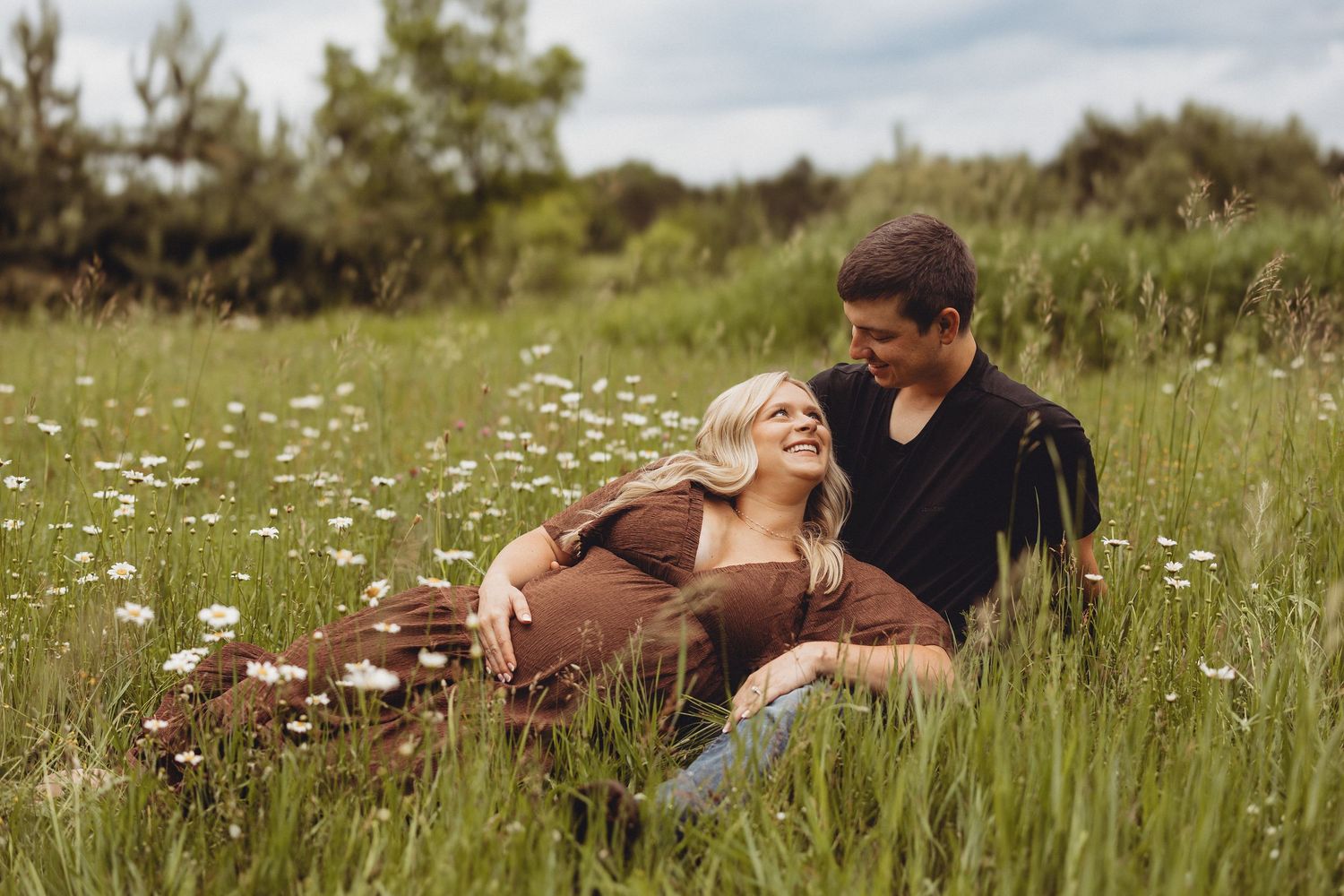 Couple sitting together in a wildflower meadow, woman leaning back smiling at man in black shirt.