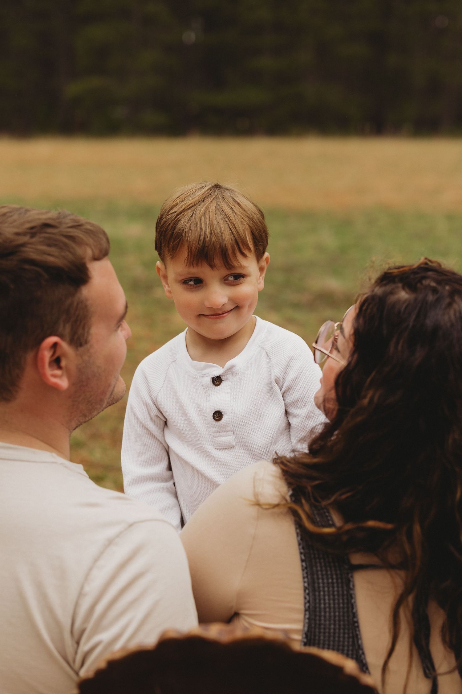 Adorable toddler boy in white henley smiling at camera while parents look on during outdoor family photo session.