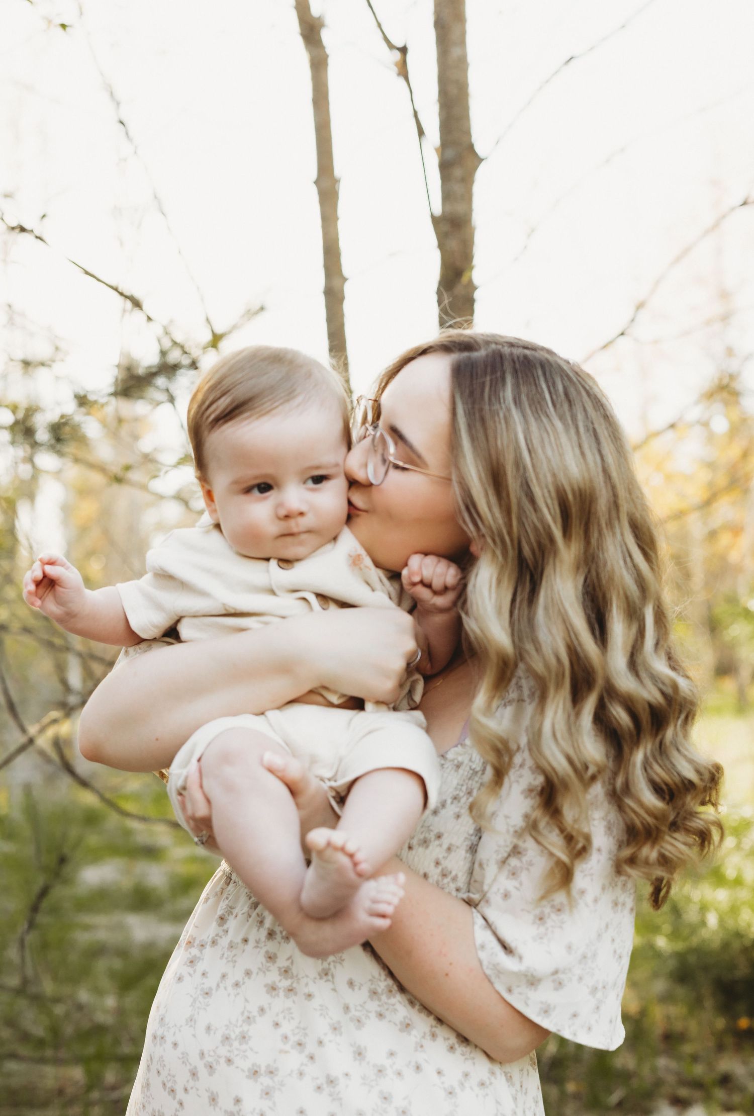 A mother with long blonde hair kisses her baby's cheek outdoors in a sunlit forest setting.