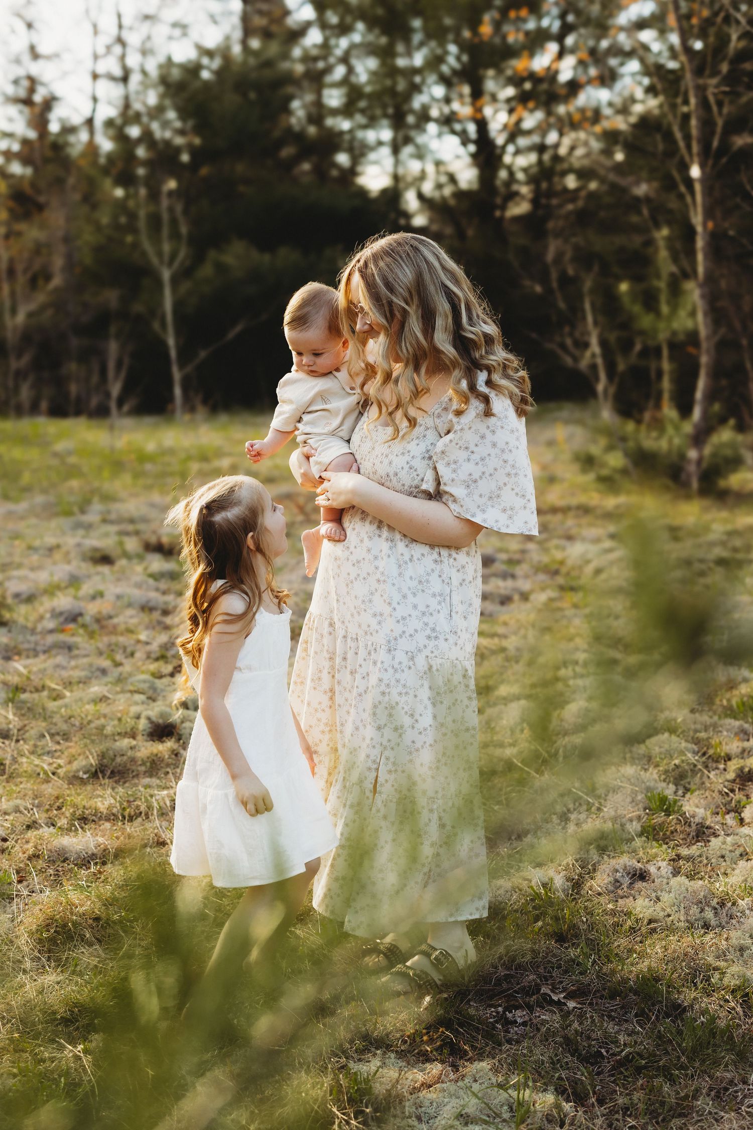 Mother in floral dress holding baby while young daughter in white dress looks up at them in sunny field.