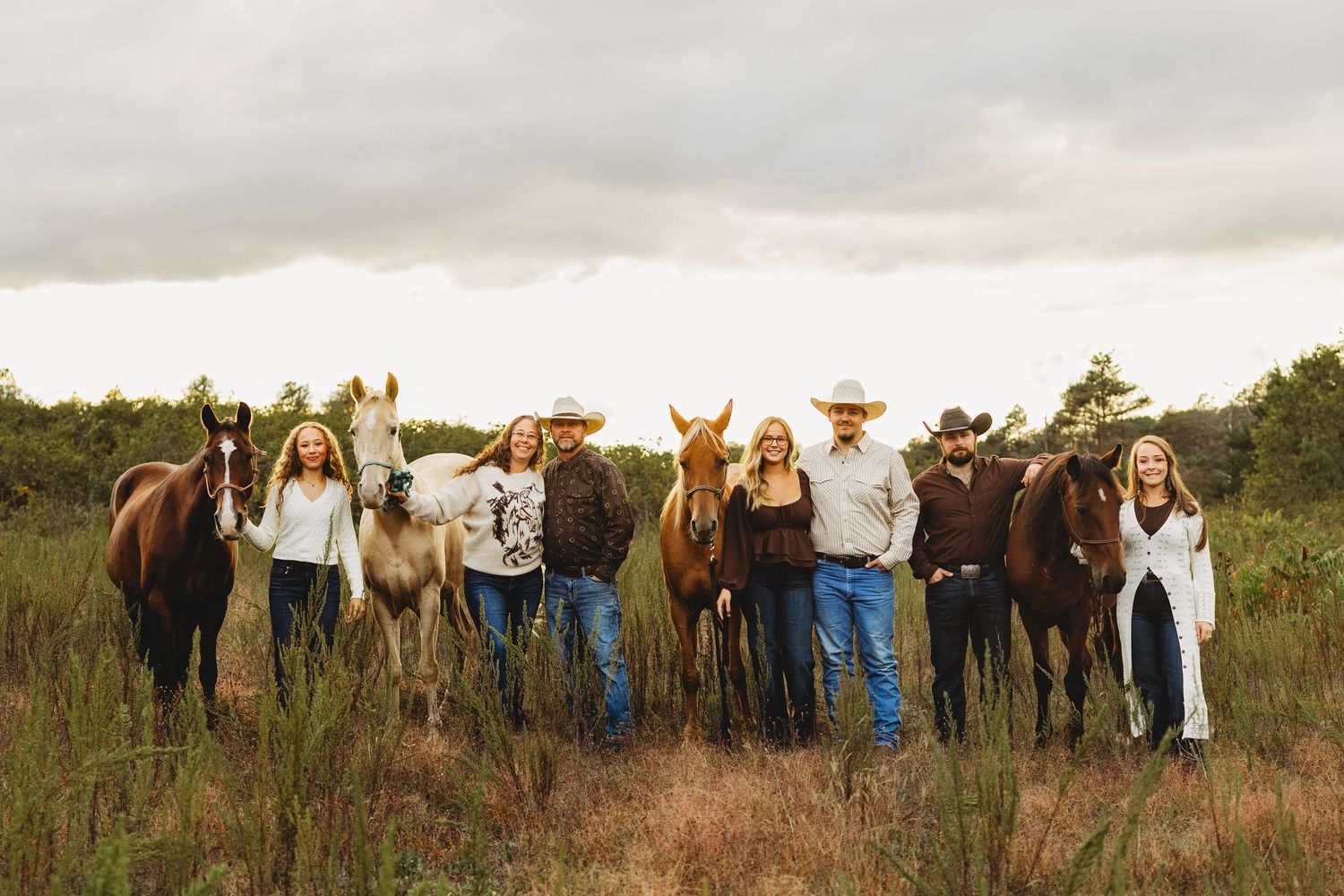 Family of seven in western attire stands in a grassy field at sunset with four horses beside them under a cloudy sky.