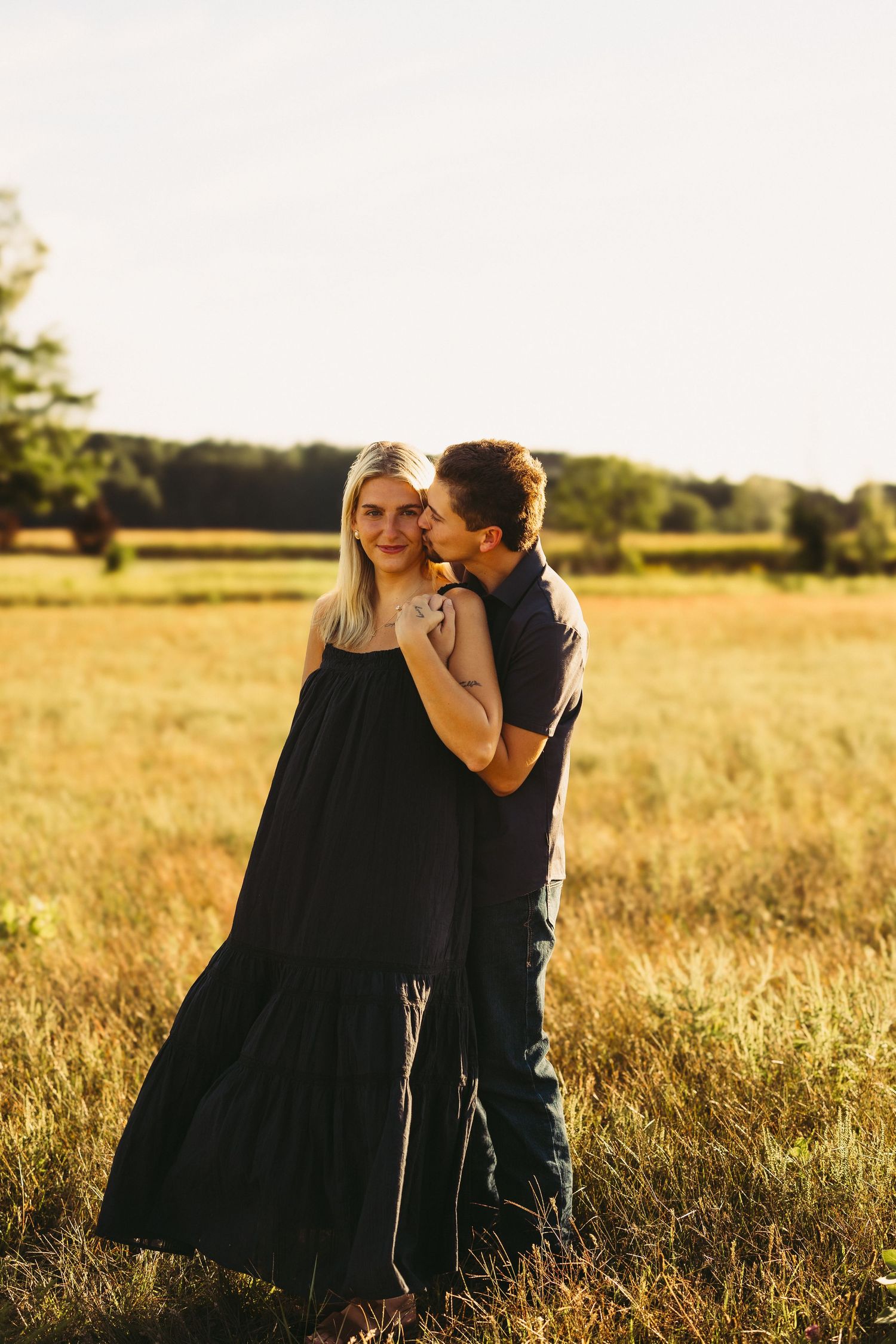 Couple embracing in a golden sunlit field as a man kisses the cheek of a smiling woman in a long black dress at sunset.