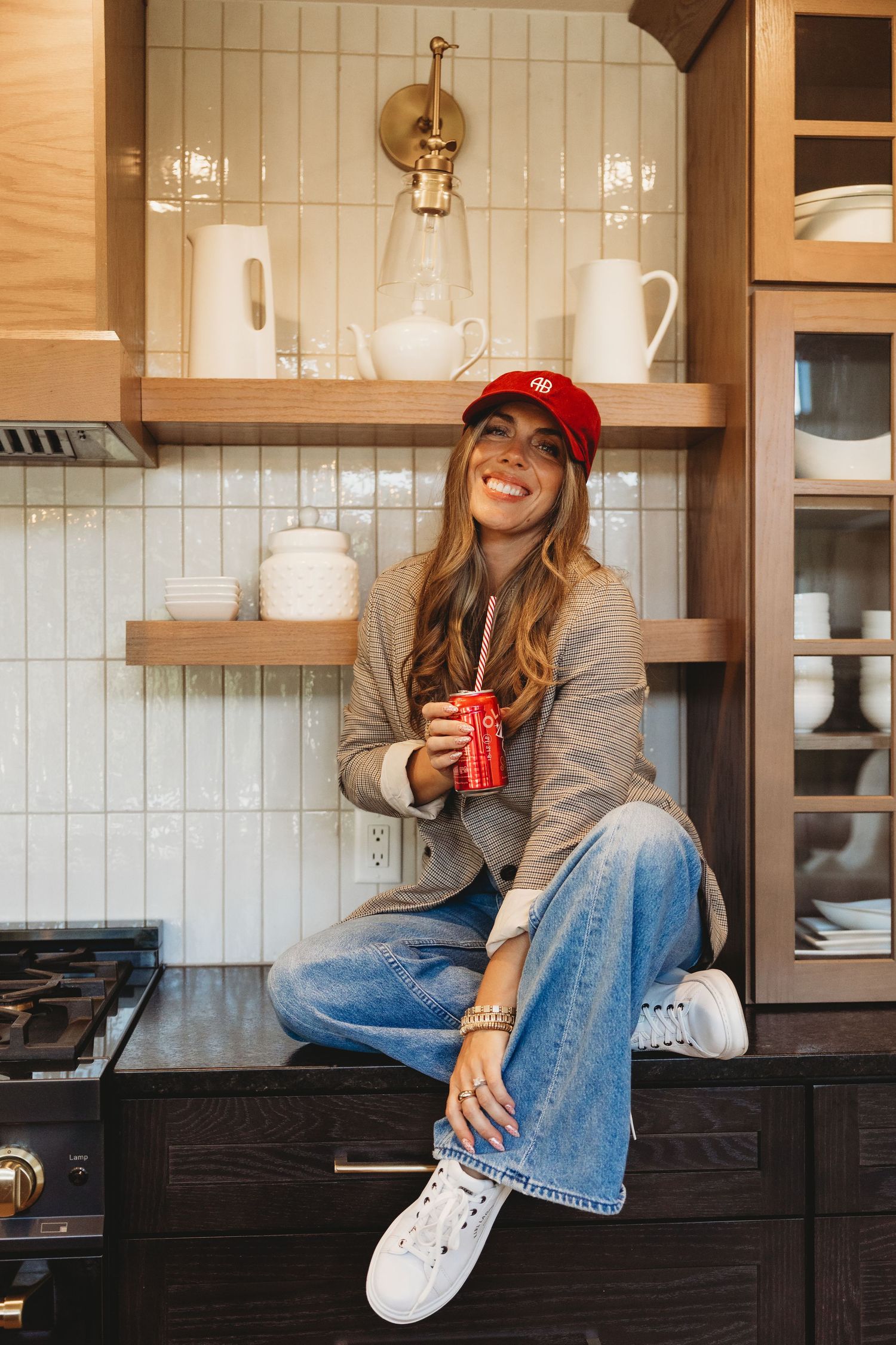 Smiling woman in a red cap, blazer and jeans sits on a kitchen counter holding a red soda can near white tile and wooden shelves.