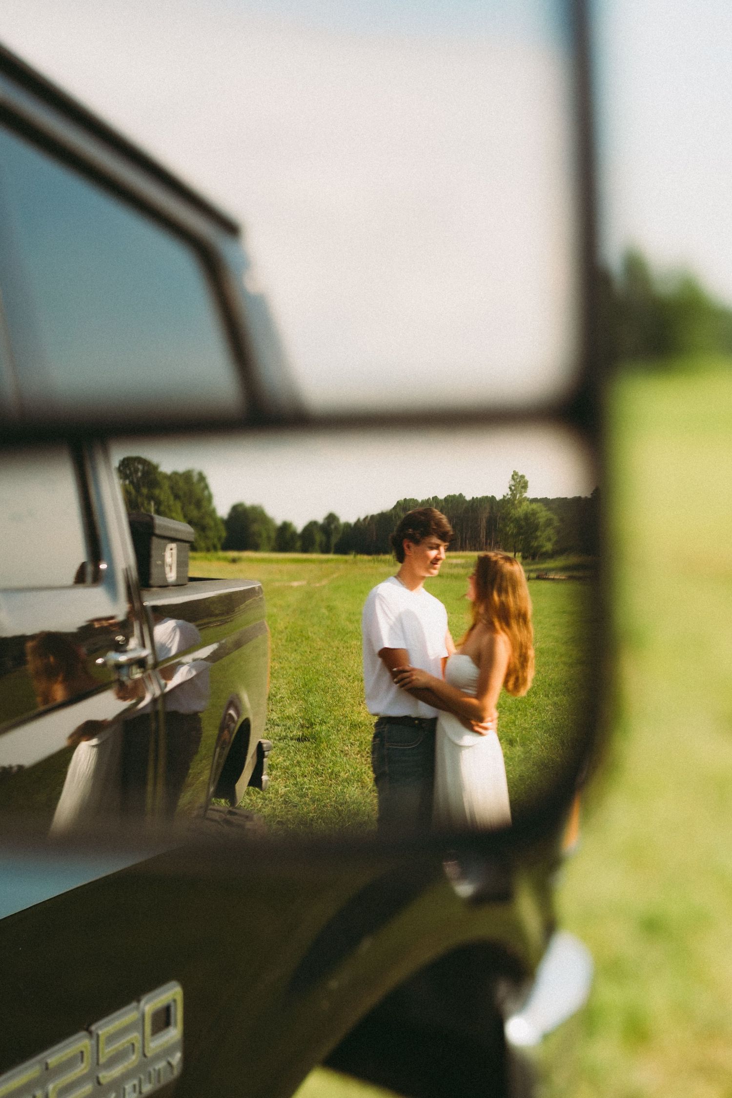Romantic couple embracing in a sunny green field, reflected in the side mirror of a vintage off-road vehicle.